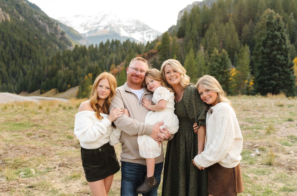A family poses for photos in a scenic mountain meadow with evergreen trees and snow-capped peaks in the background.