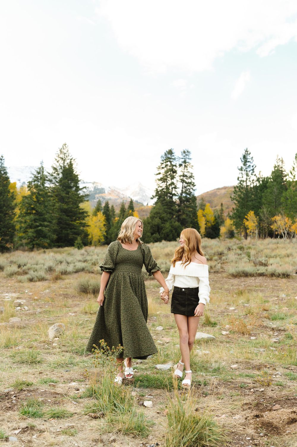 Two friends walk together through a mountain meadow surrounded by evergreen trees and autumn foliage.