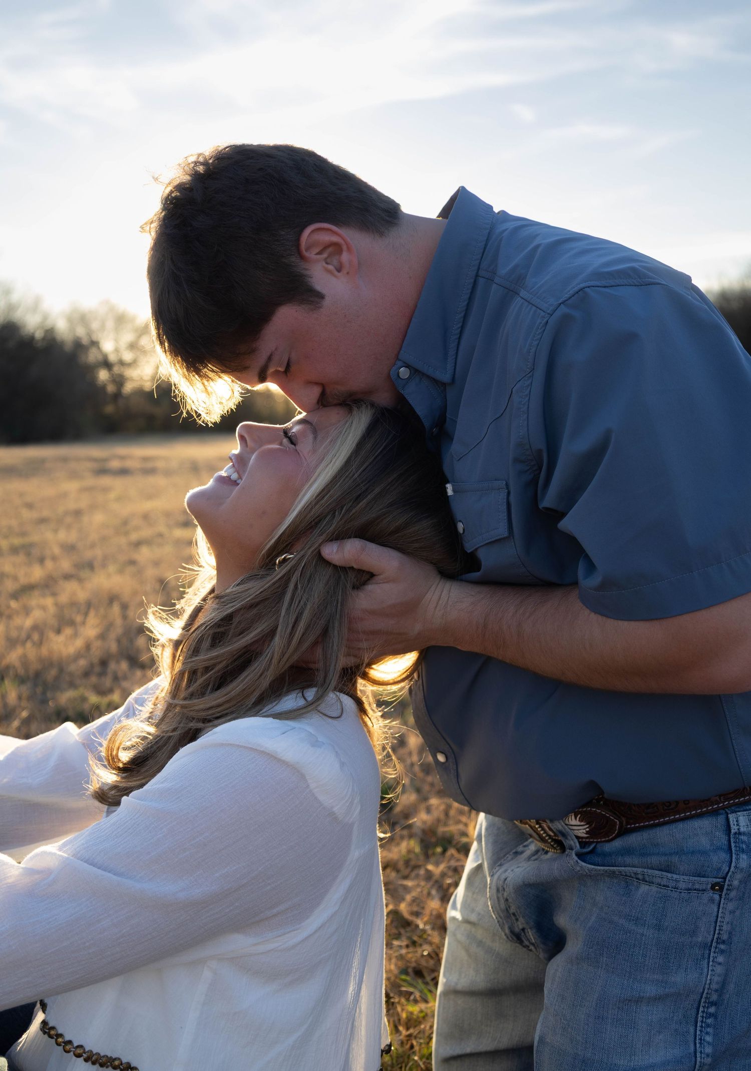 Romantic couple sharing a kiss in an open field at golden hour during an outdoor engagement session.