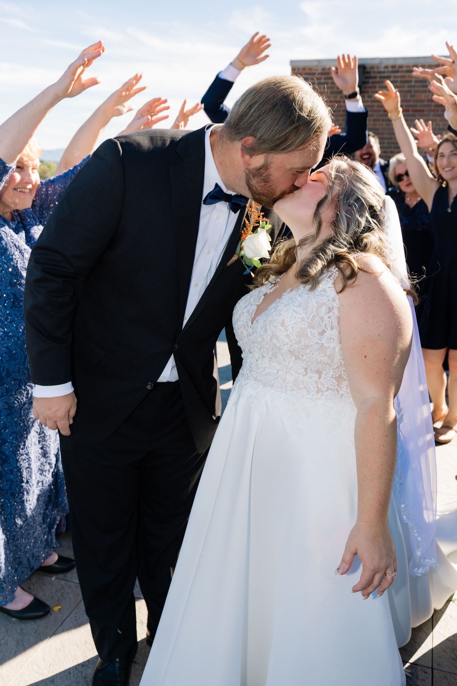 A newlywed couple shares a kiss while wedding guests cheer and raise their hands in celebration behind them.