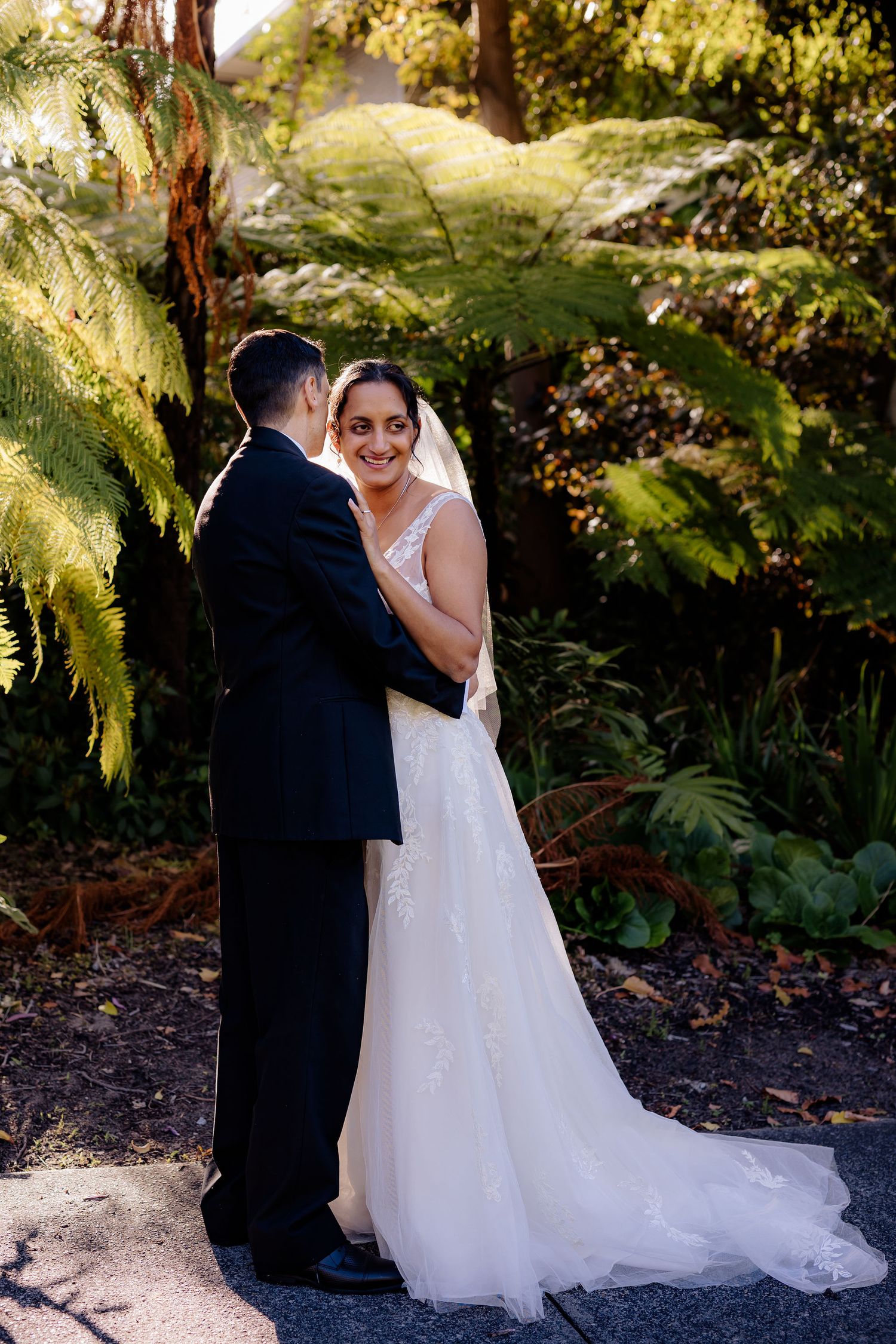 A couple in formal wedding attire embraces in a lush garden setting with ferns and dappled sunlight.