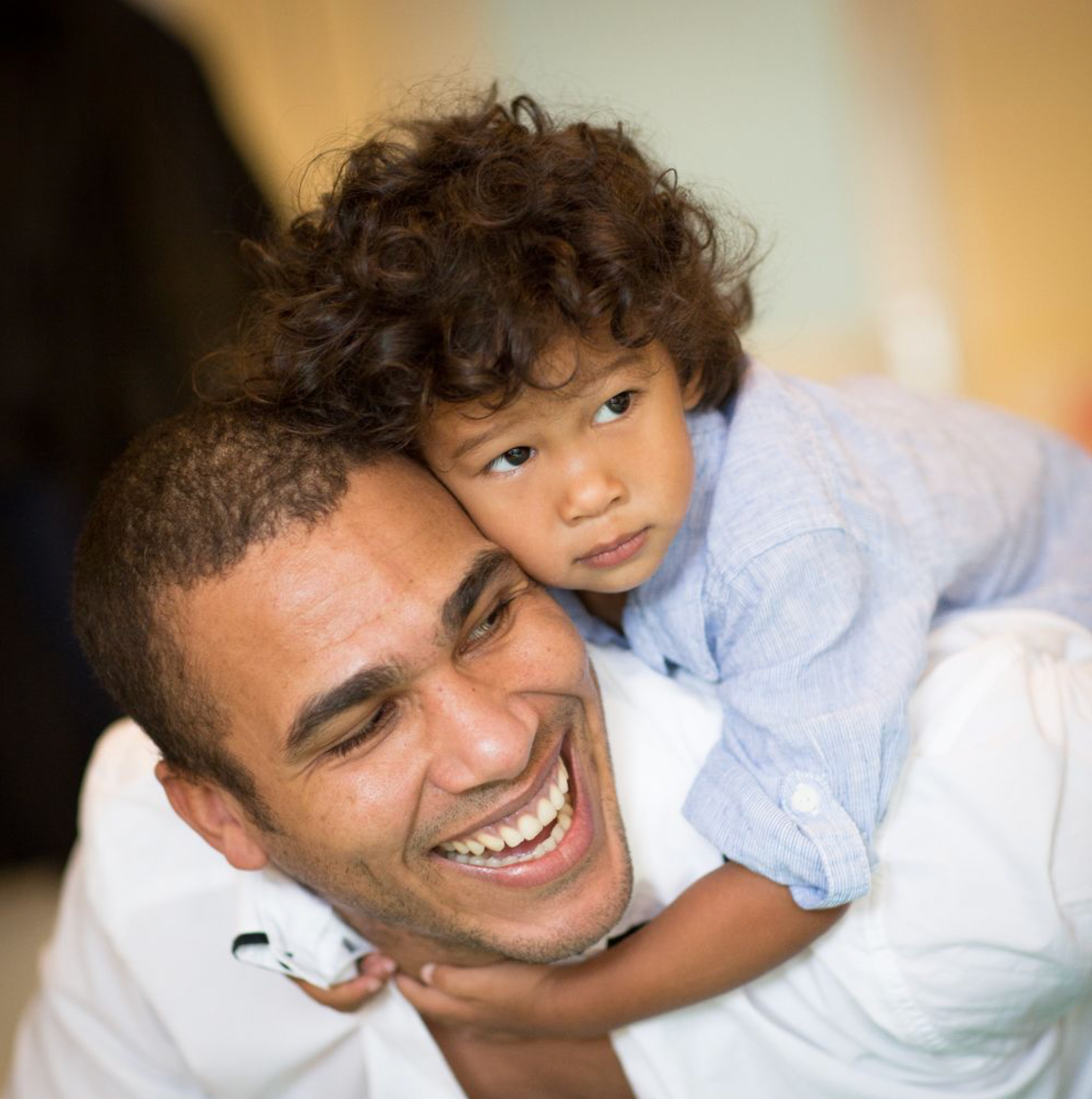 A laughing man with a curly-haired toddler riding on his back, both smiling happily.