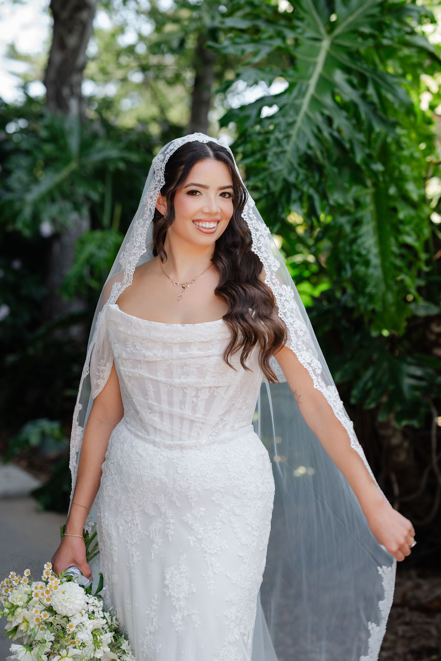 A bride in a white lace wedding dress with long sleeves holds a delicate white and green floral bouquet in a garden setting.