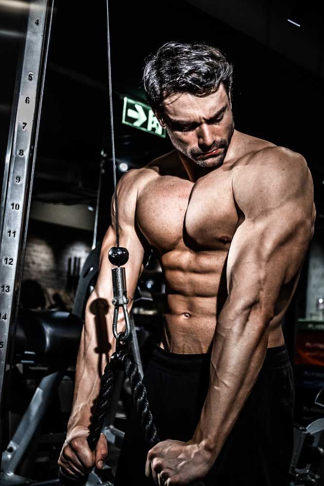 A muscular shirtless man working out on a cable machine in a dark gym.