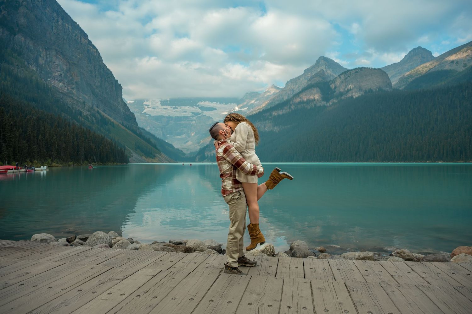 A dog plays and poses on a wooden dock overlooking a serene mountain lake surrounded by towering peaks.