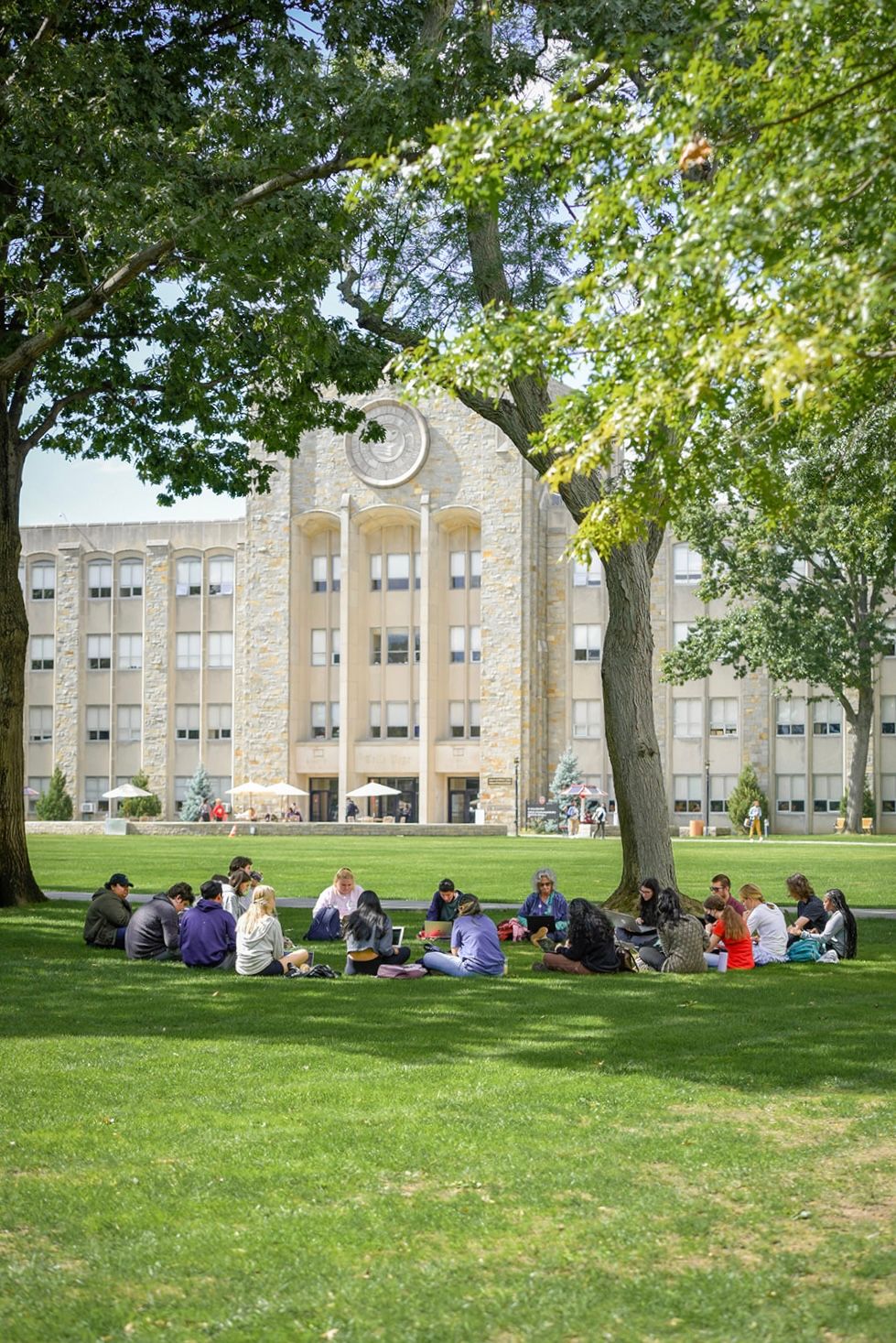 Students relax on a grassy lawn in front of a large stone academic building on a sunny afternoon.