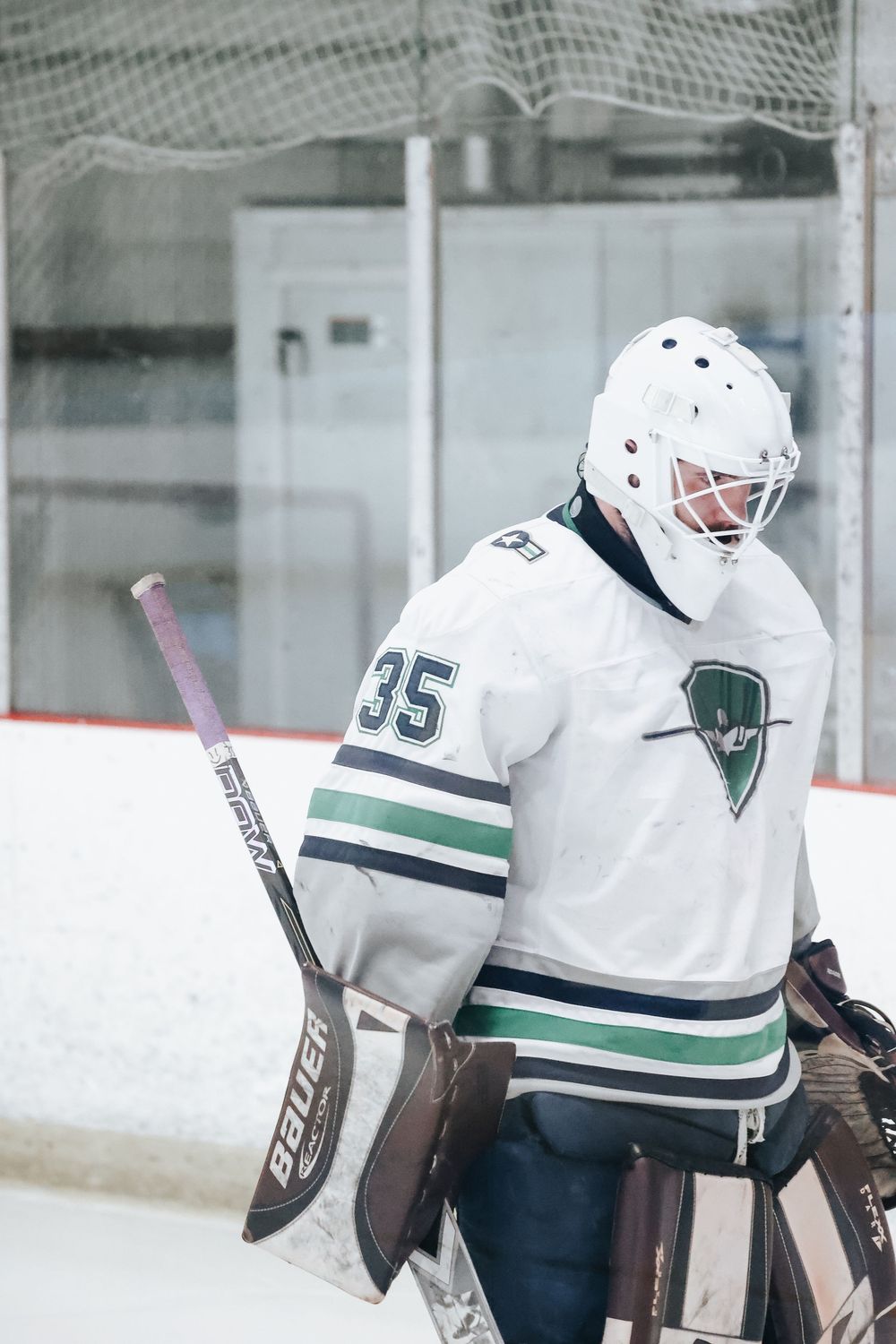 Hockey goalies in white jerseys with green stripes line up along the ice rink boards during practice.