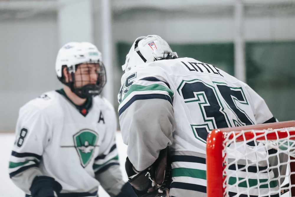 Hockey players in white and green uniforms compete near the red goal post during game action.