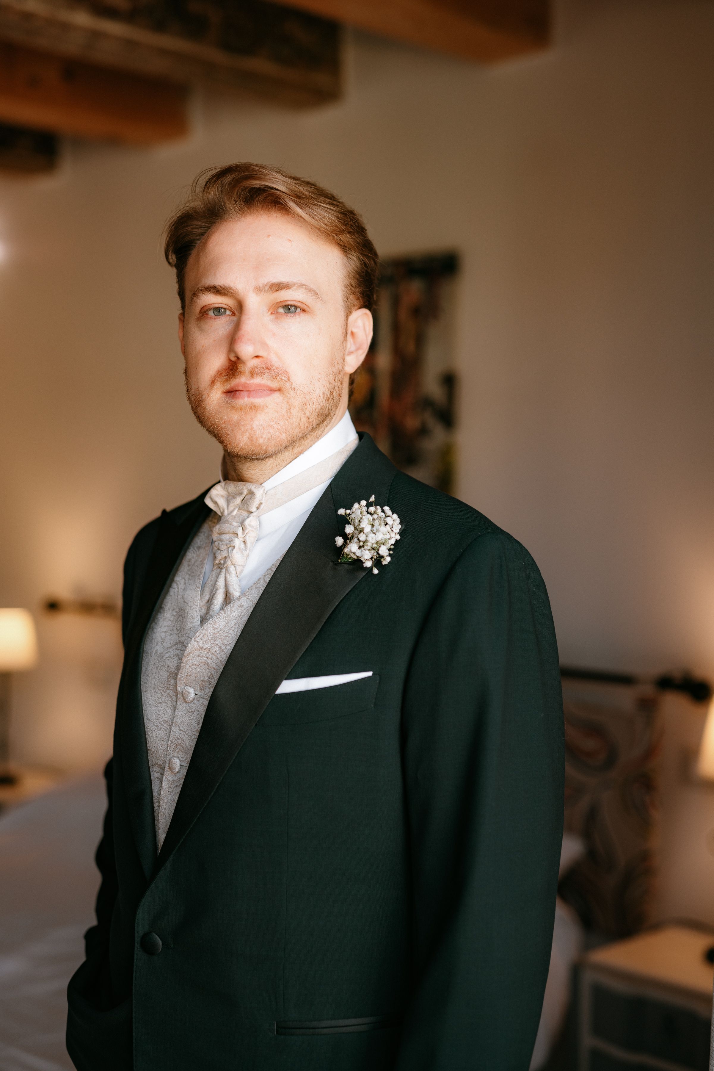 A groom wearing a classic black tuxedo with a white boutonniere poses in a rustic indoor setting.