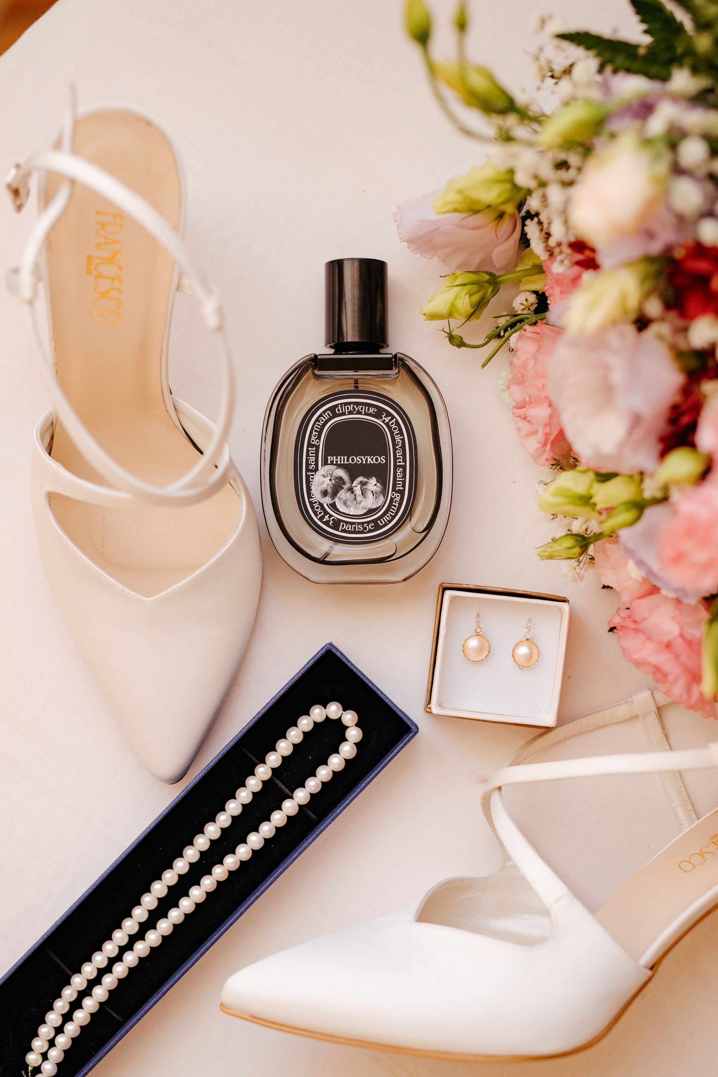 Elegant bridal accessories including white heels, pearl jewelry, and perfume arranged with pink flowers and navy boxes.