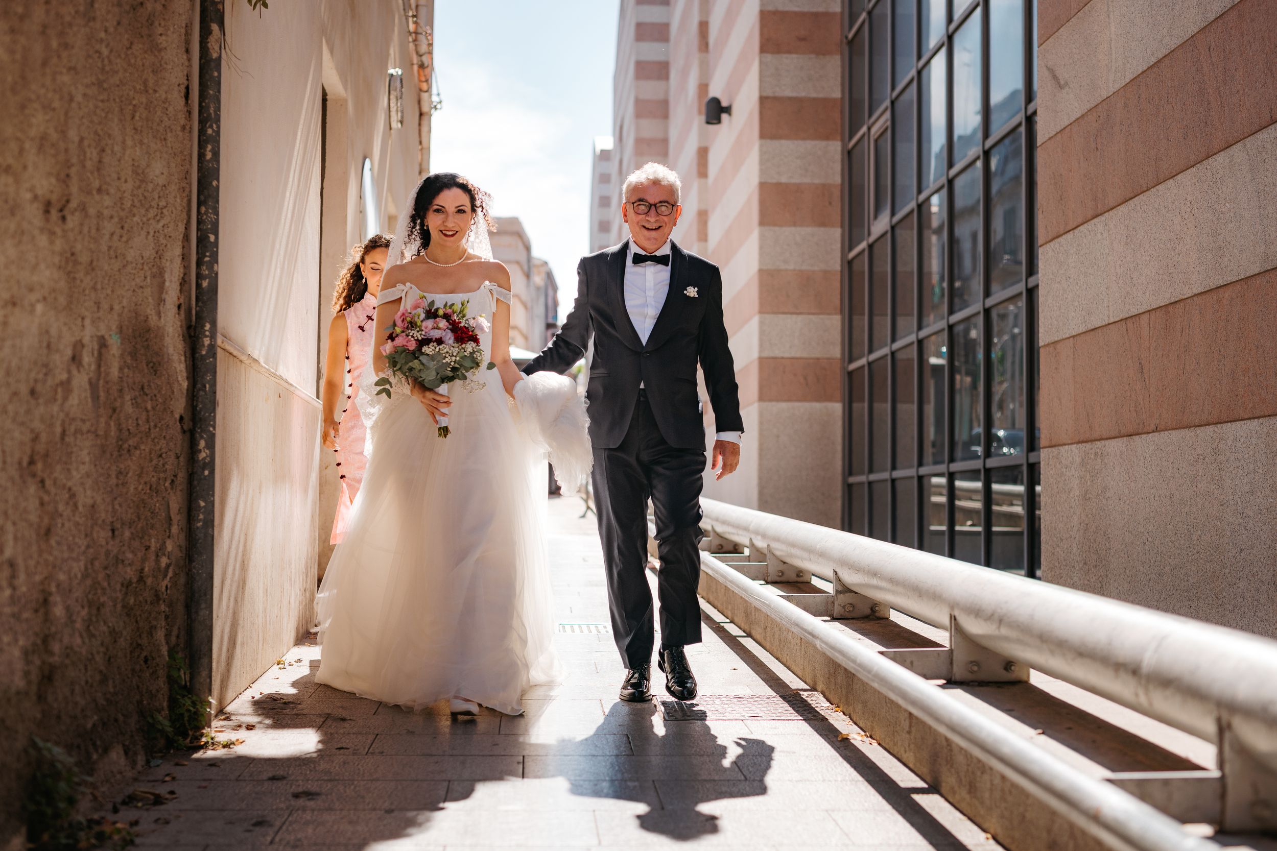 Couple in formal wedding attire walking through a sunlit stone corridor with dramatic shadows and light.