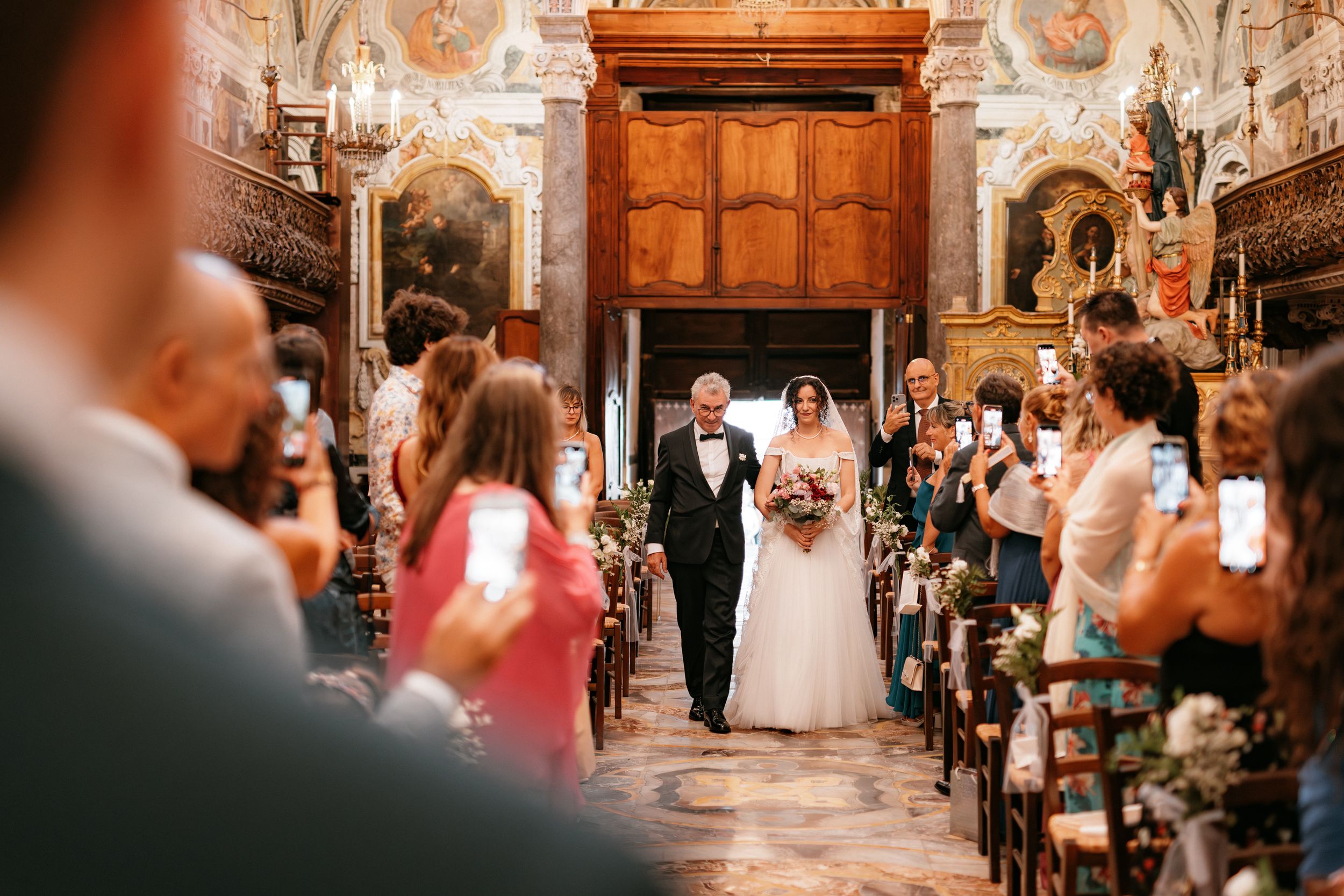 Wedding guests gather in an ornate church interior with warm lighting and decorative frescoes on the walls.