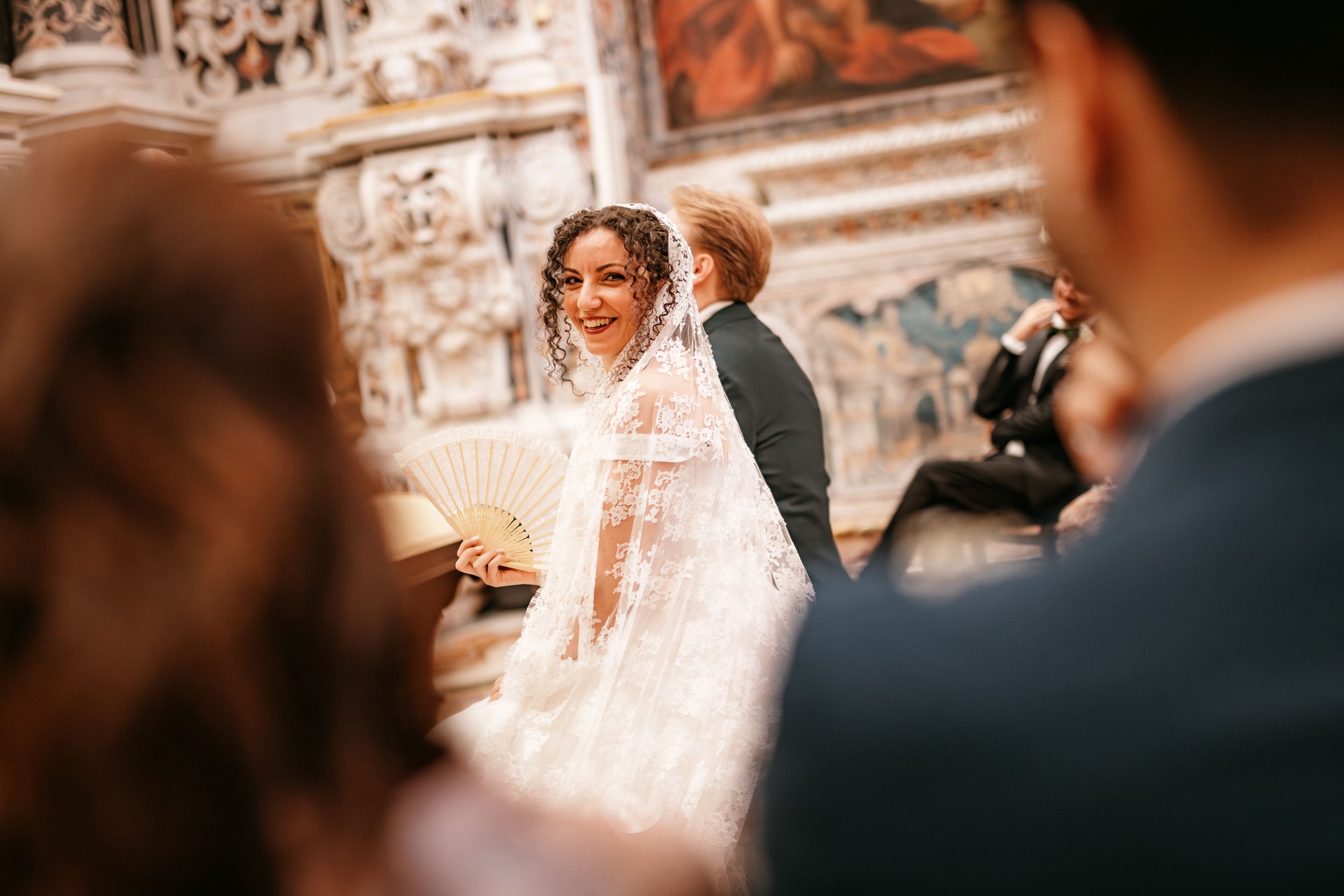 Wedding ceremony guests gathered in a historic church with ornate stone architecture and warm lighting.