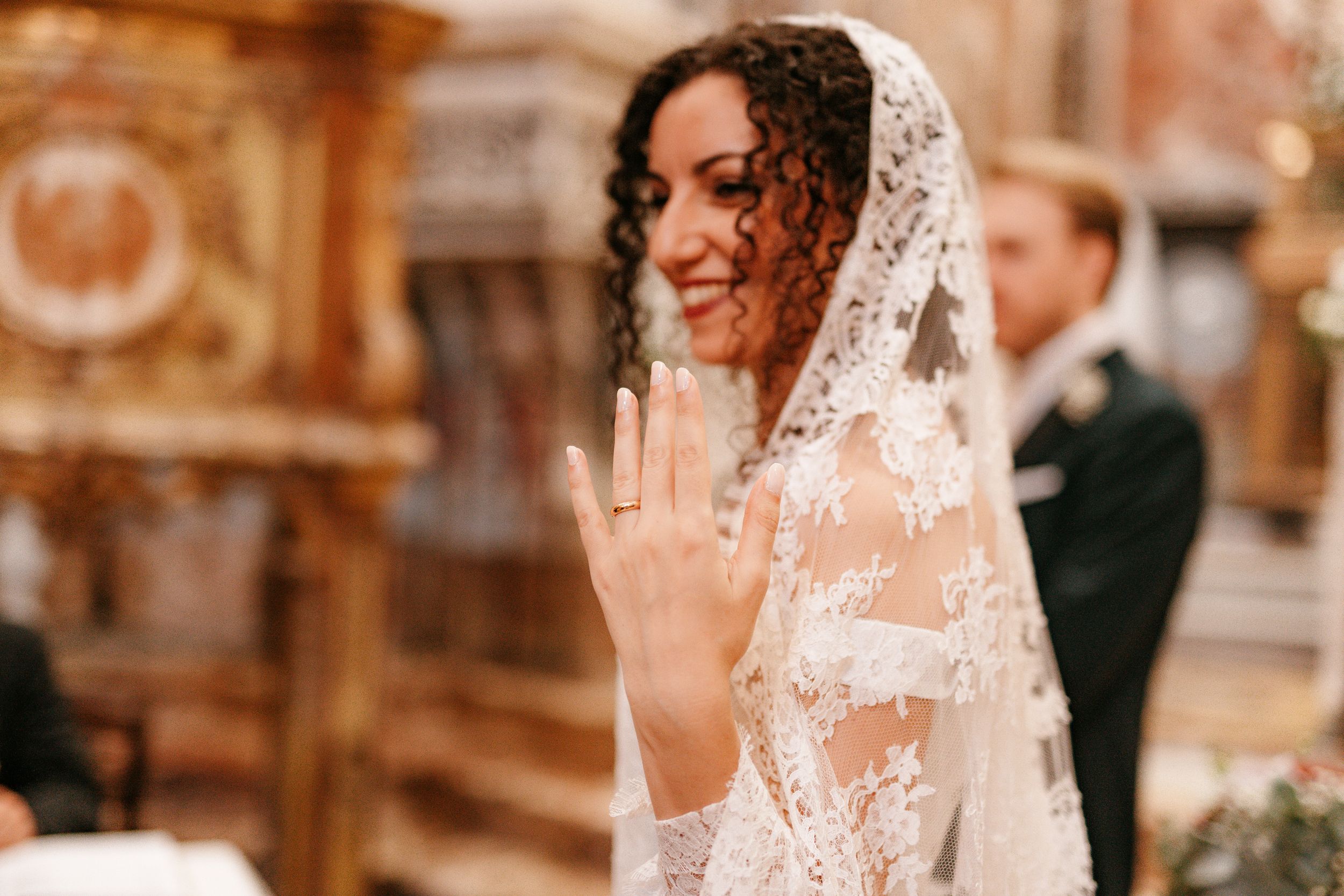 A bride wearing an elegant lace veil smiles while showing her wedding ring in a church setting.