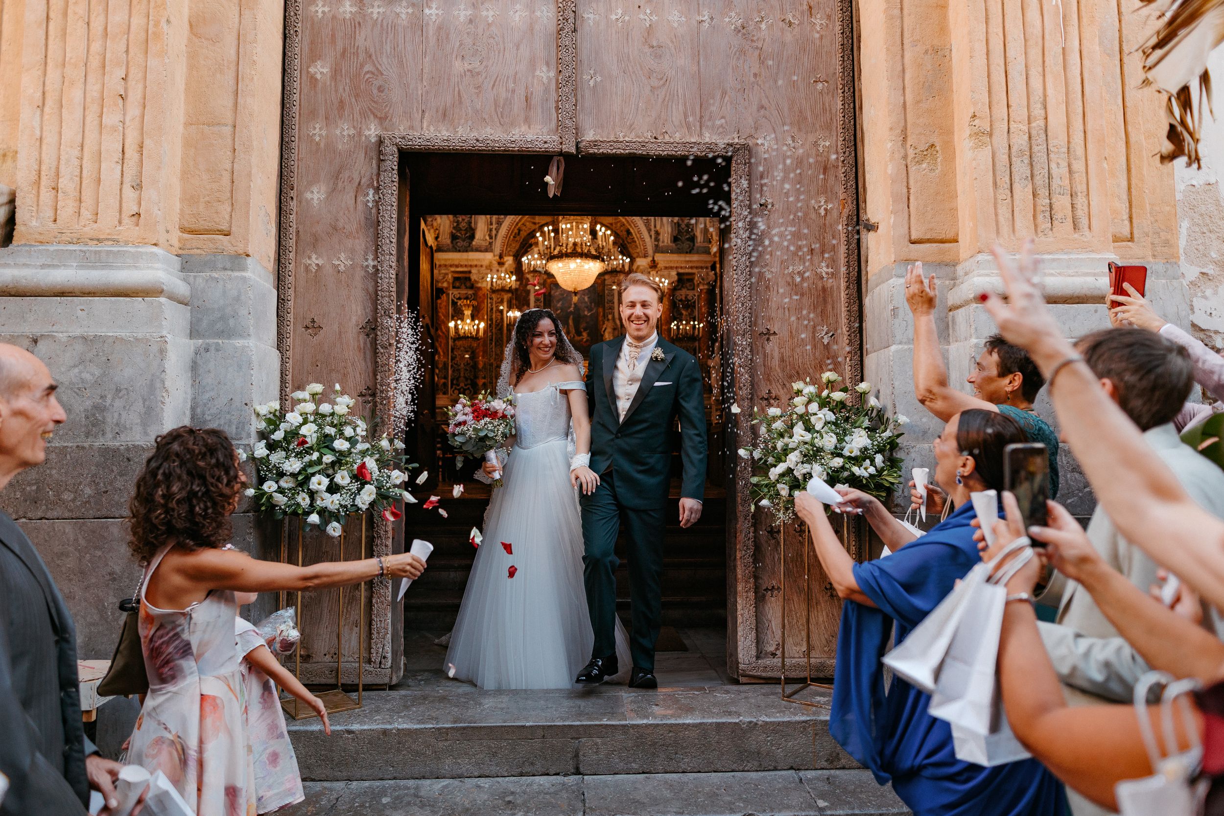 Wedding celebration outside historic church entrance with floral arrangements and guests throwing confetti.