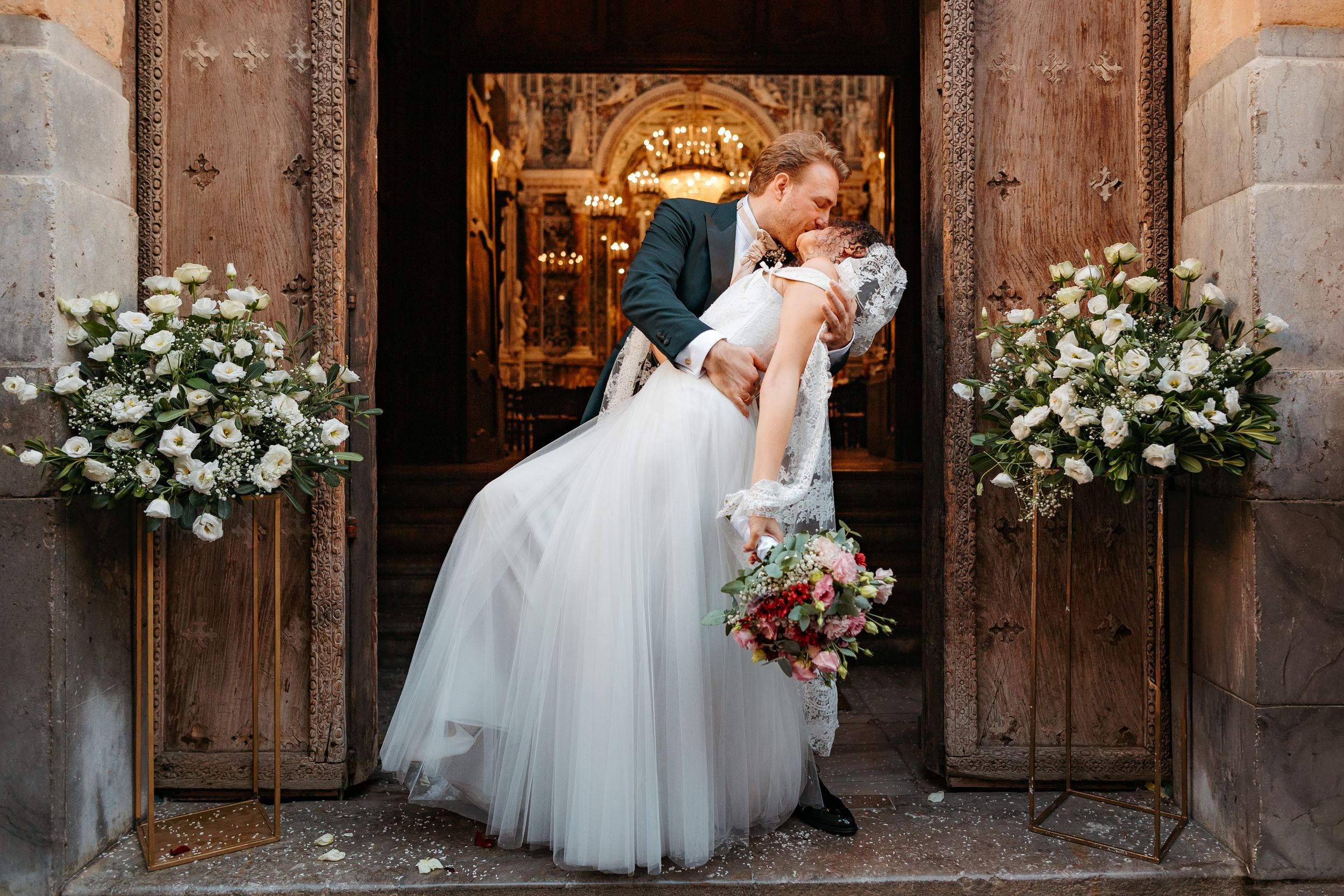 A romantic wedding couple shares a kiss in front of wooden church doors decorated with white floral arrangements.