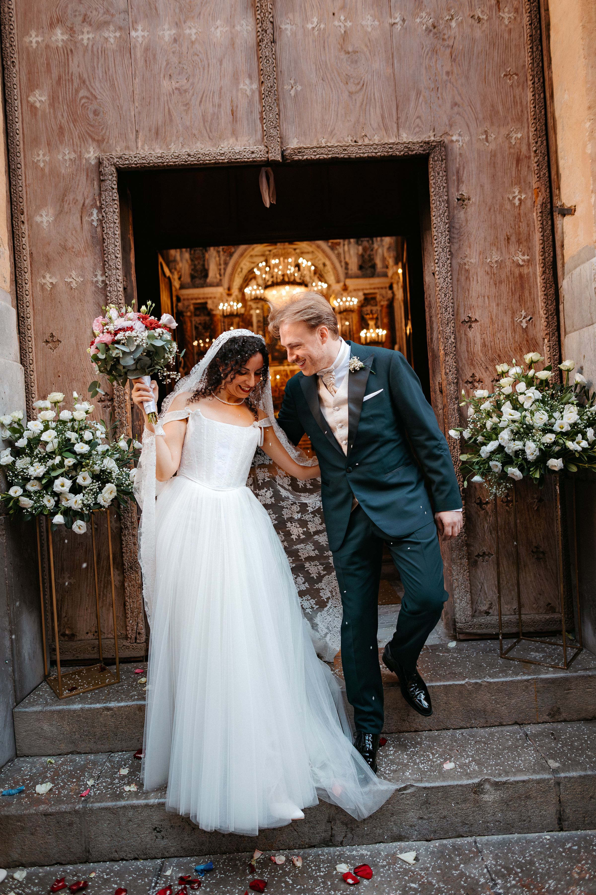 A wedding couple shares a playful moment peeking through rustic wooden doors decorated with white floral arrangements.