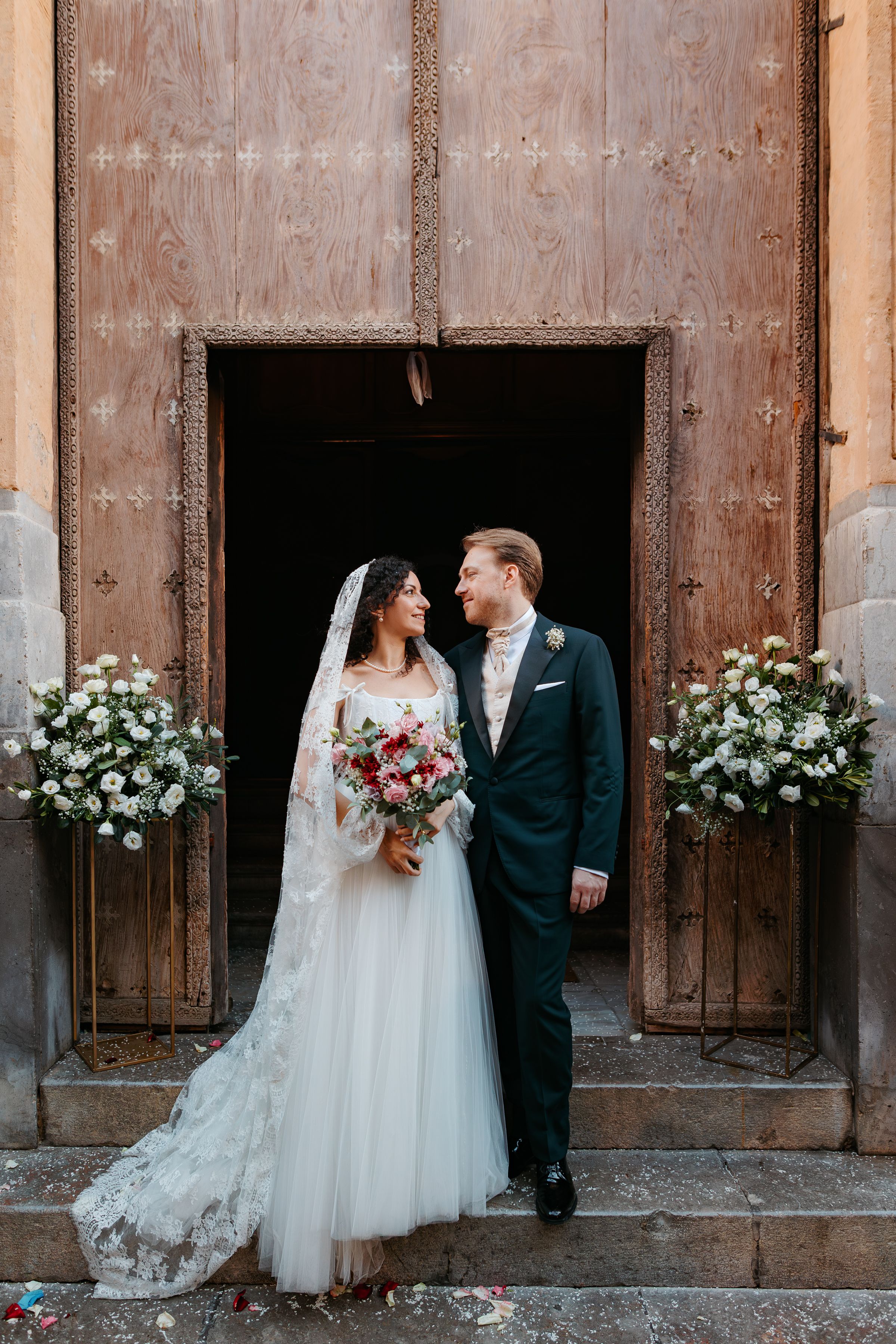 A couple stands in a rustic wooden doorway decorated with white floral arrangements on their wedding day.