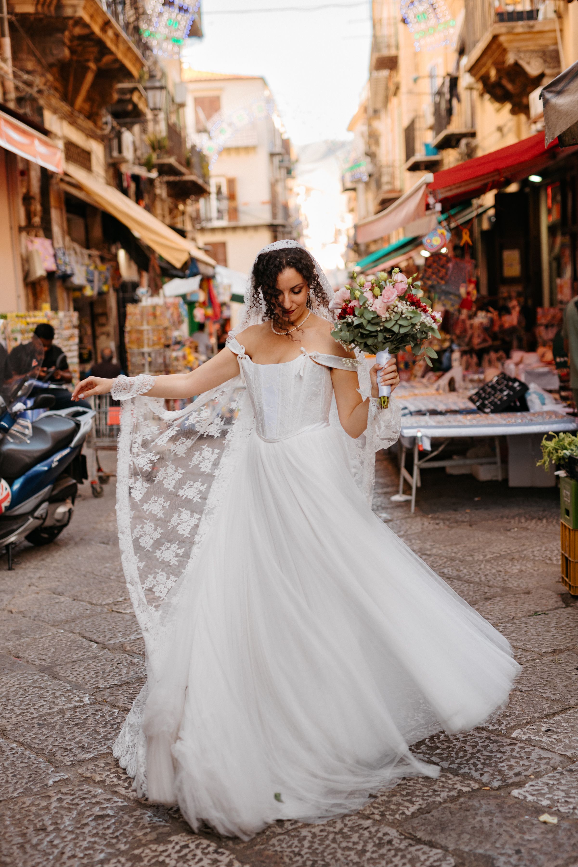 A bride in a flowing white lace wedding dress twirls gracefully on a charming cobblestone street in Italy.