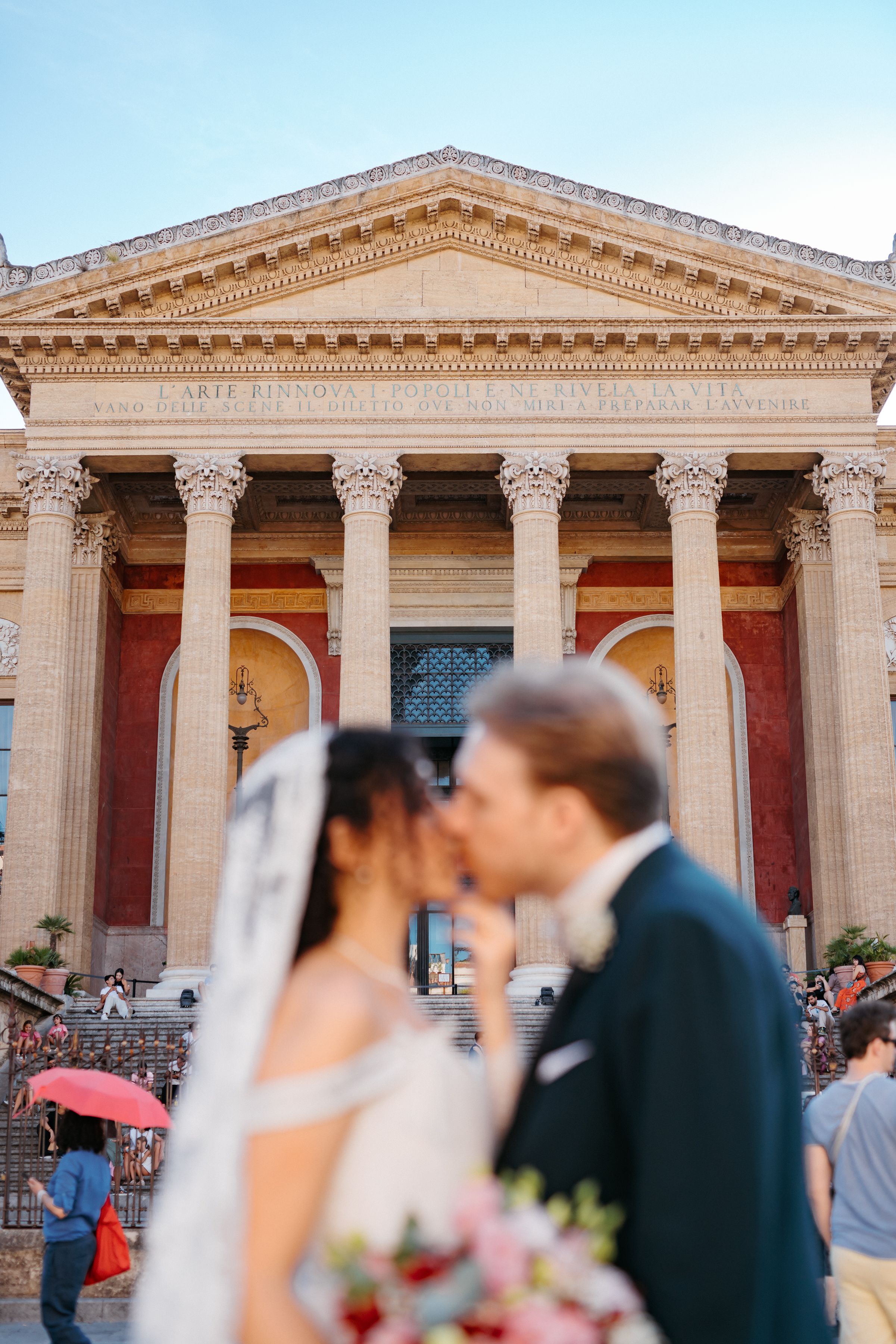A wedding couple shares a romantic moment outside a grand neoclassical building with ornate columns and architecture.