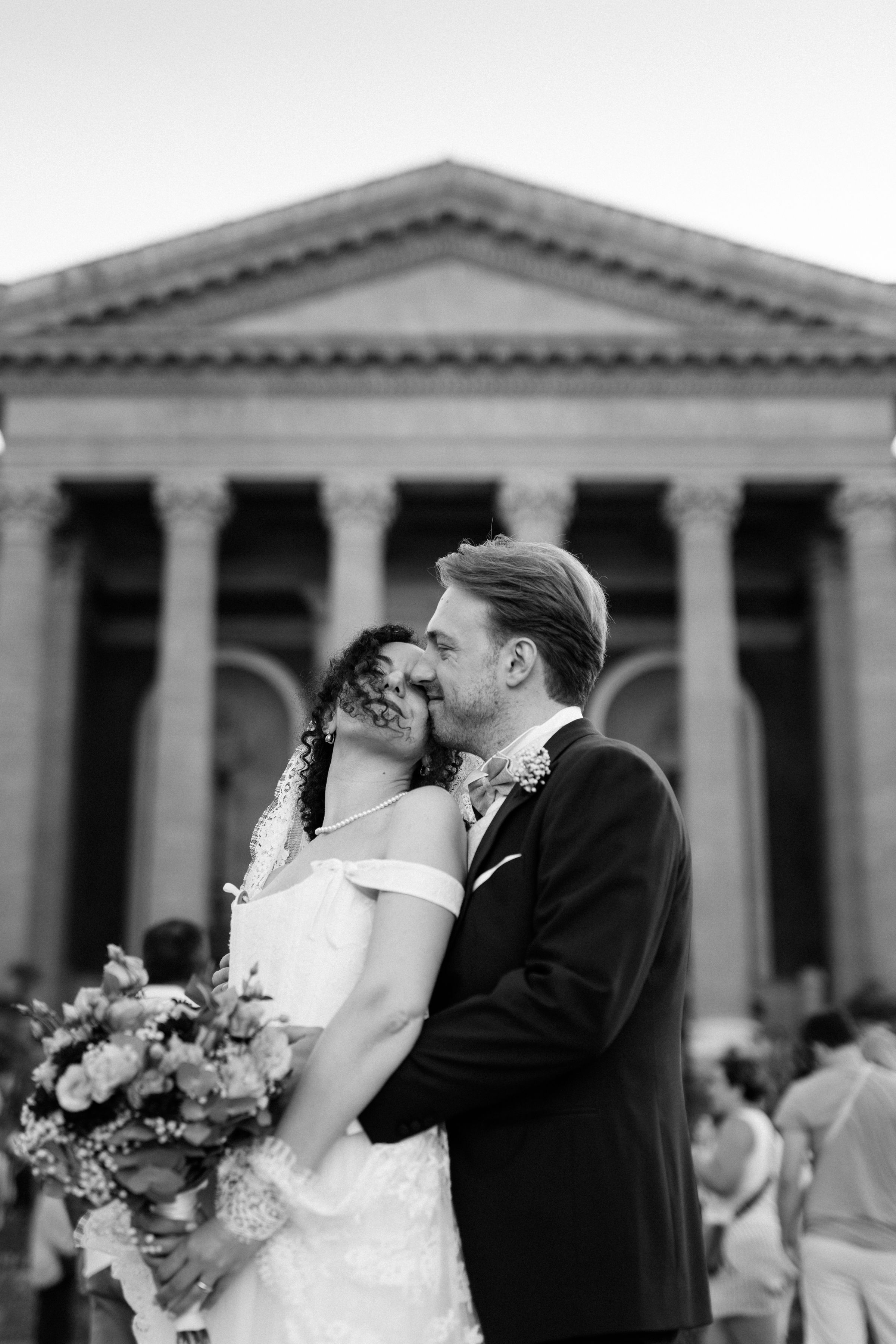 A romantic wedding kiss moment captured in front of a grand columned building with classic architecture.