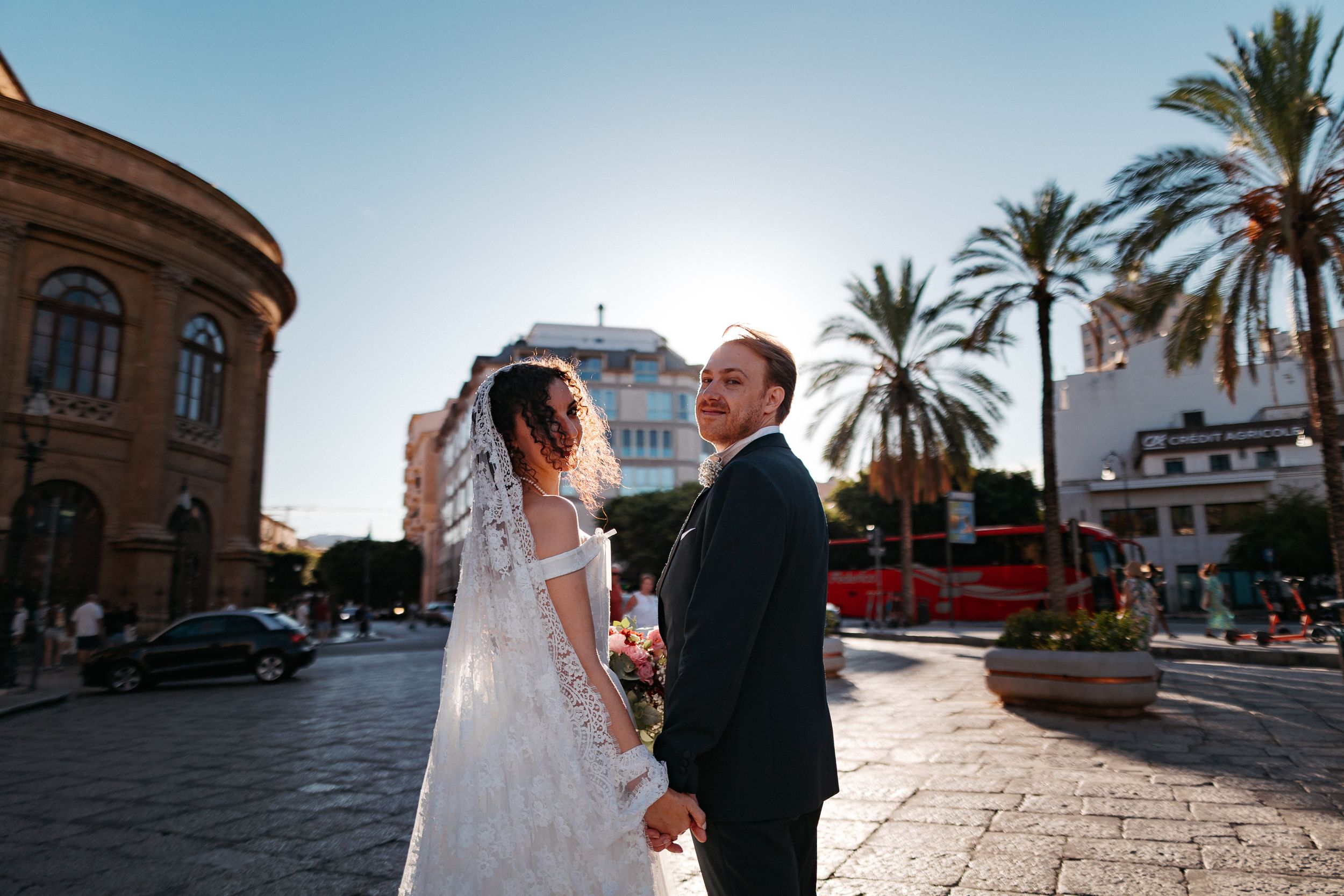 Elegant wedding couple poses against Mediterranean architecture with palm trees and historic buildings in the background.