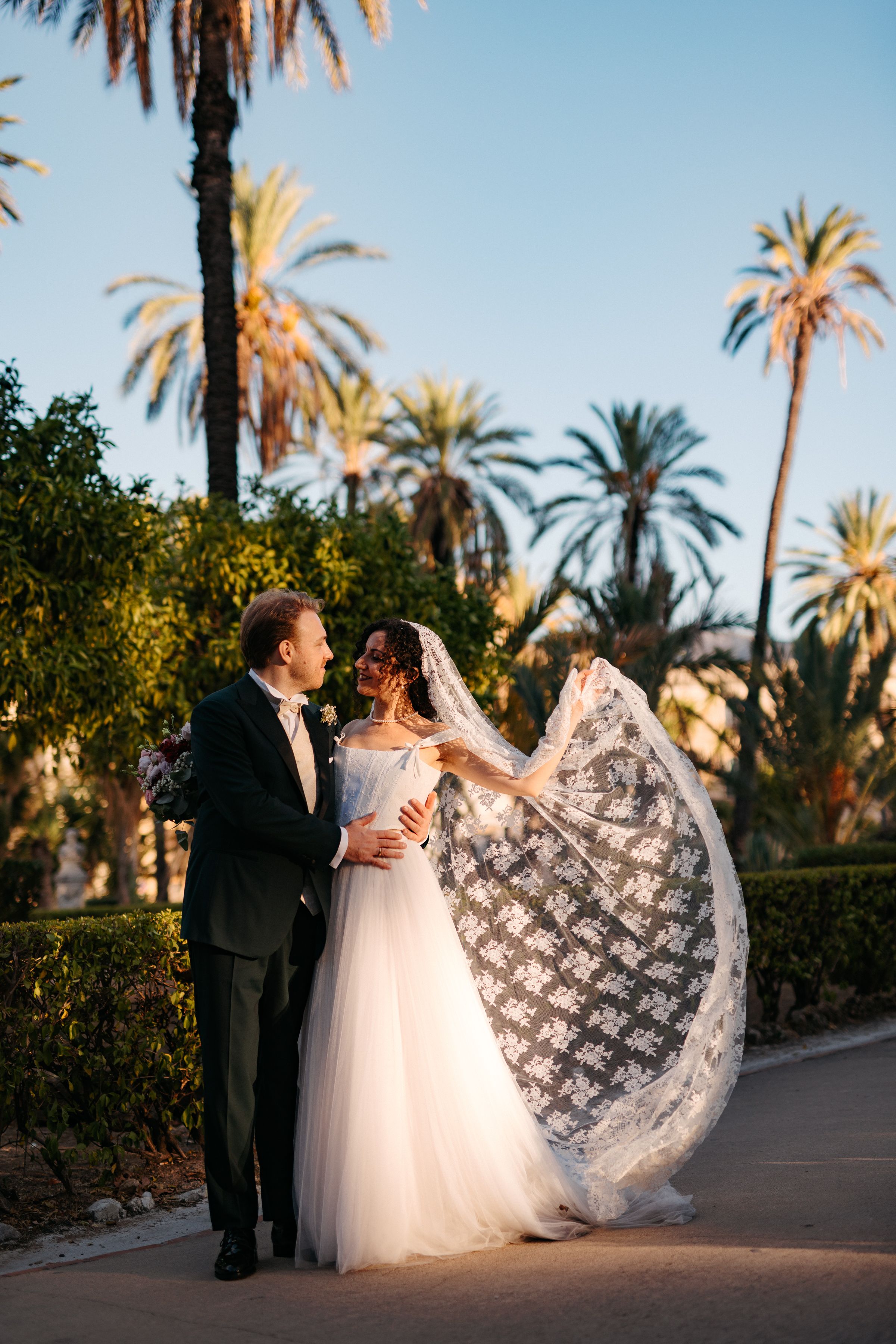 A romantic wedding couple dances together on a palm tree-lined path at sunset.