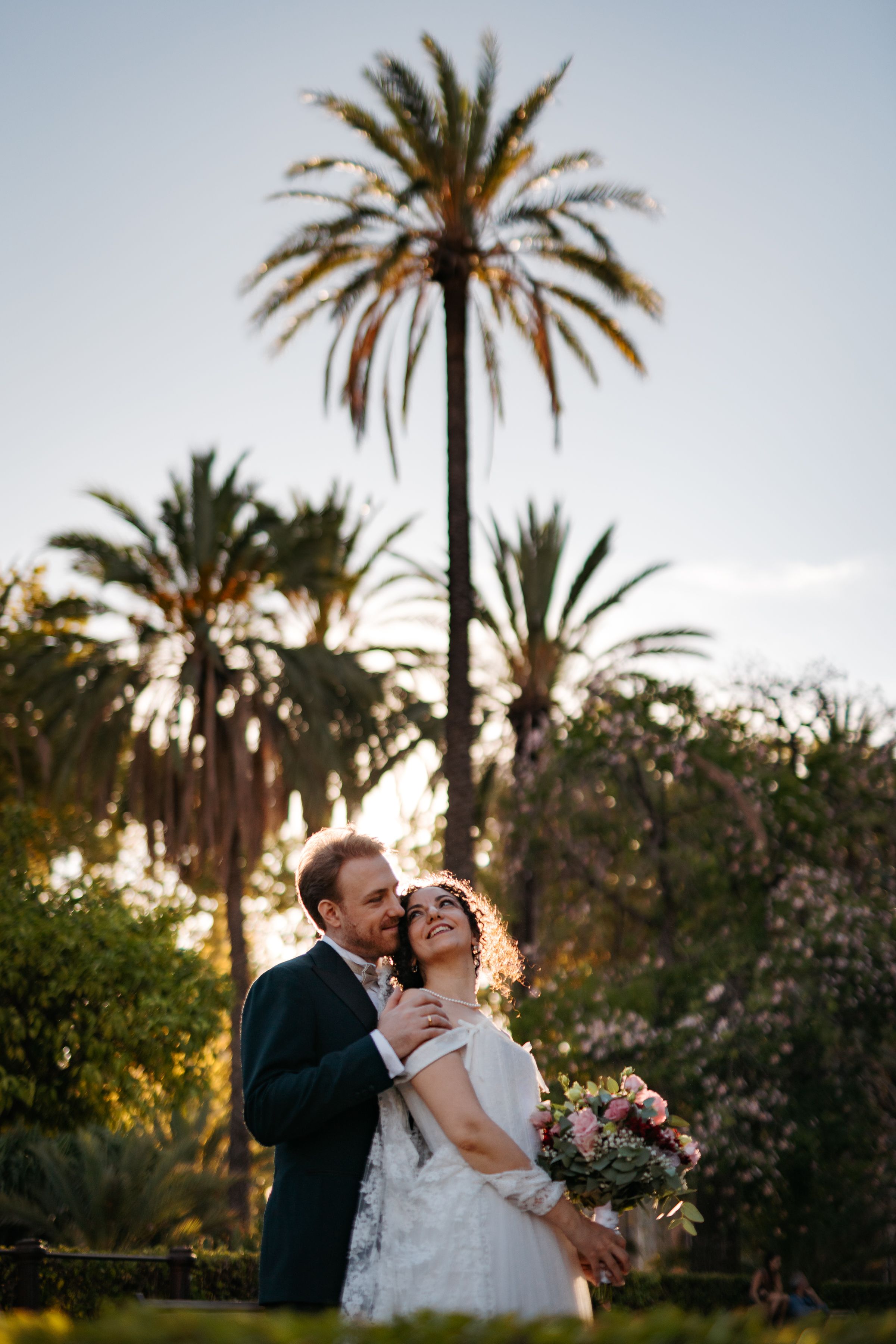Couple embraces in a romantic garden setting with palm trees and blooming flowers at sunset.