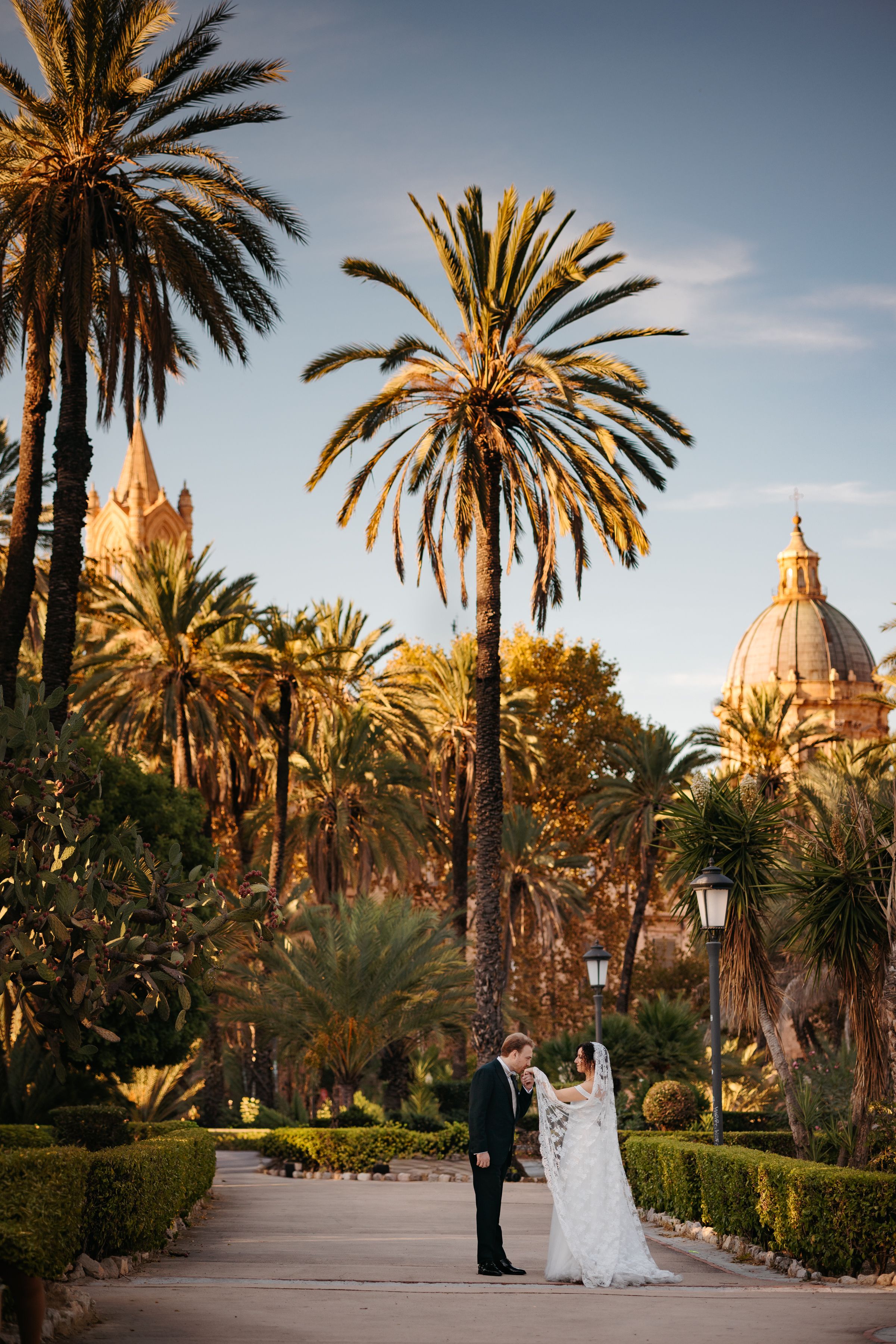 Elegant couple poses in a lush garden path lined with palm trees and a domed building in the background at sunset.