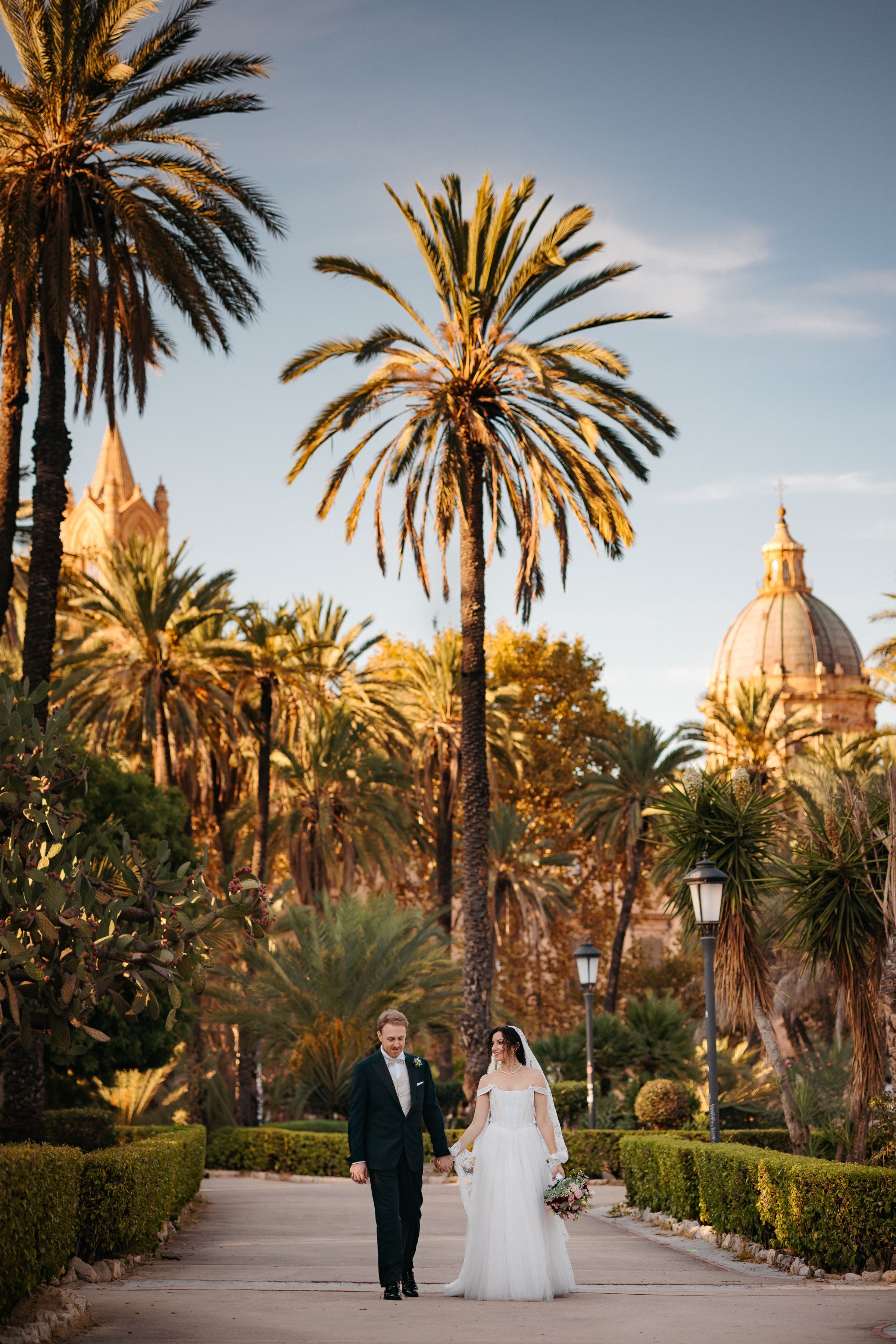 A couple walks down a palm-lined path with a grand domed building in the background during golden hour.