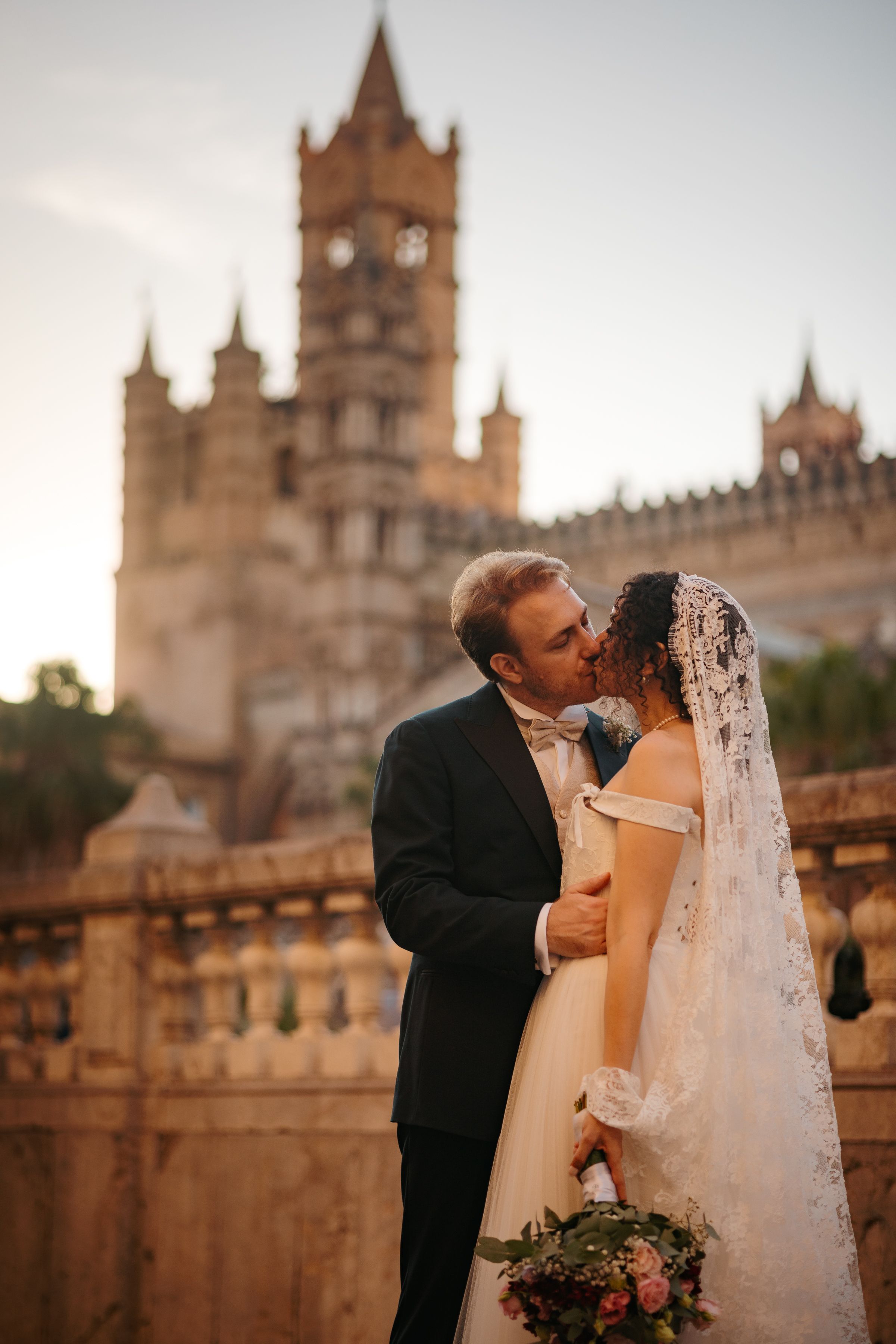 A romantic wedding couple shares intimate kisses against the backdrop of a grand gothic cathedral at sunset.
