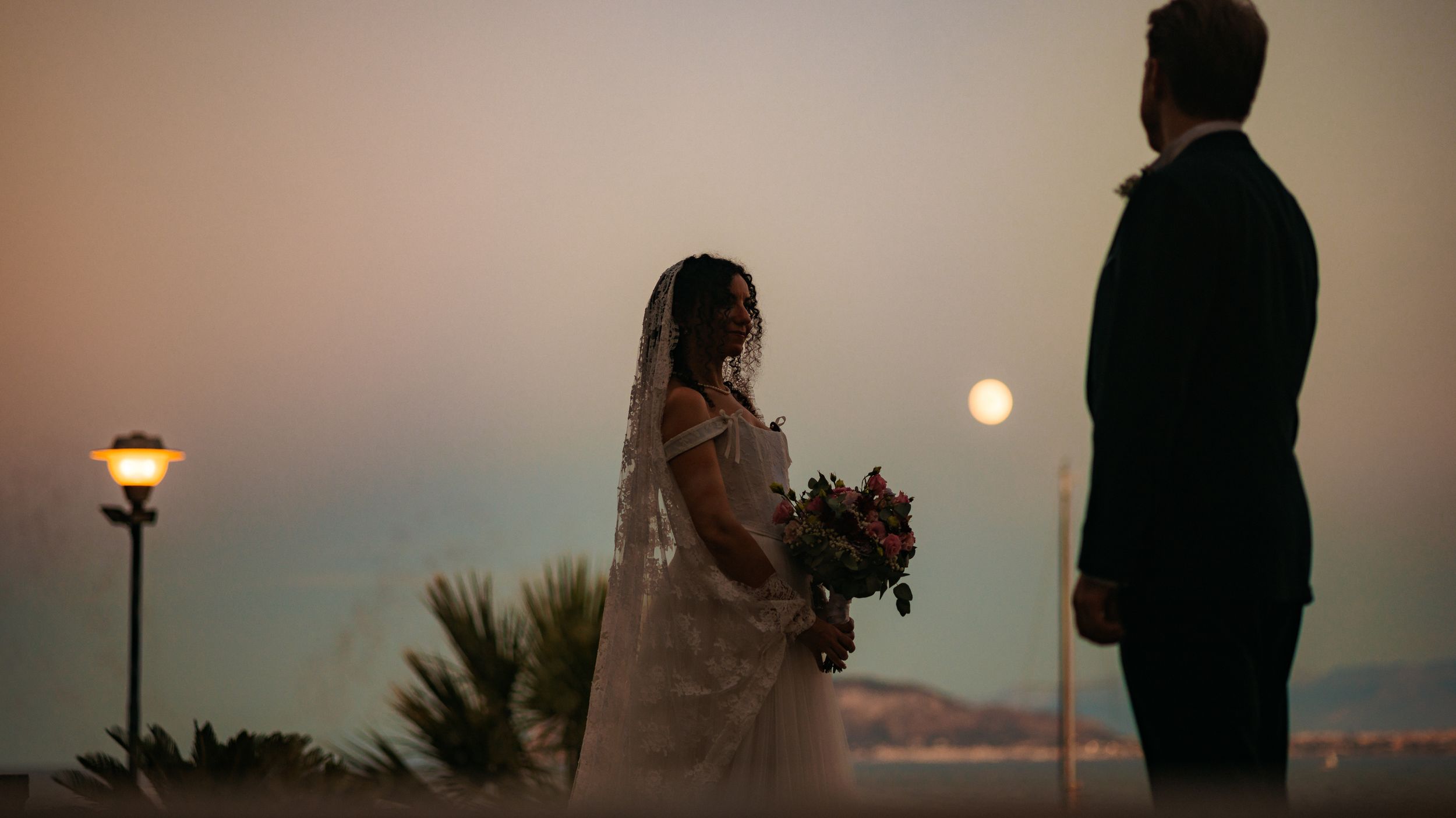 A silhouetted couple stands together against a sunset sky with palm trees and warm lighting in the background.