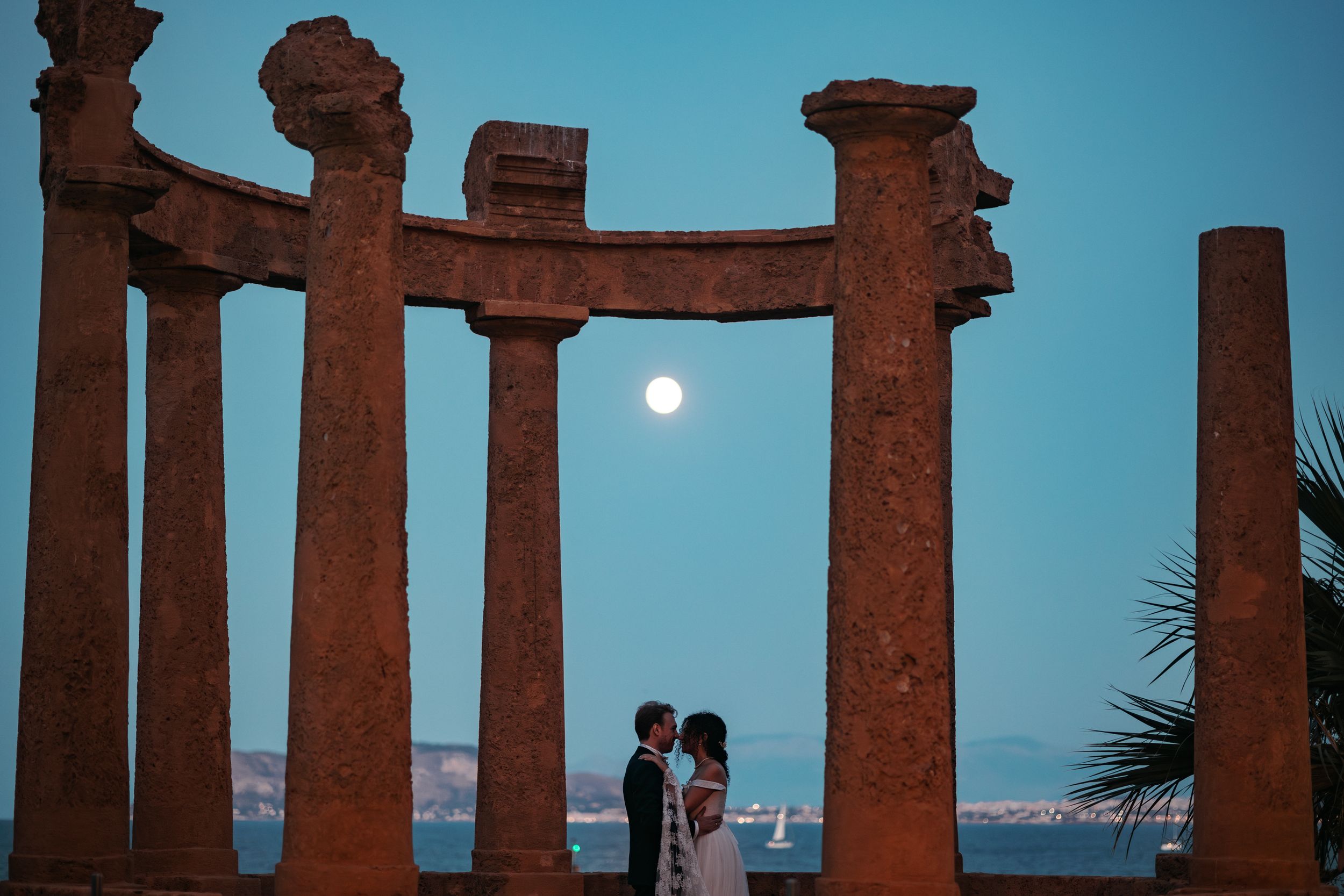 Ancient Roman columns silhouetted against a blue sky with palm trees and distant mountains at sunset.