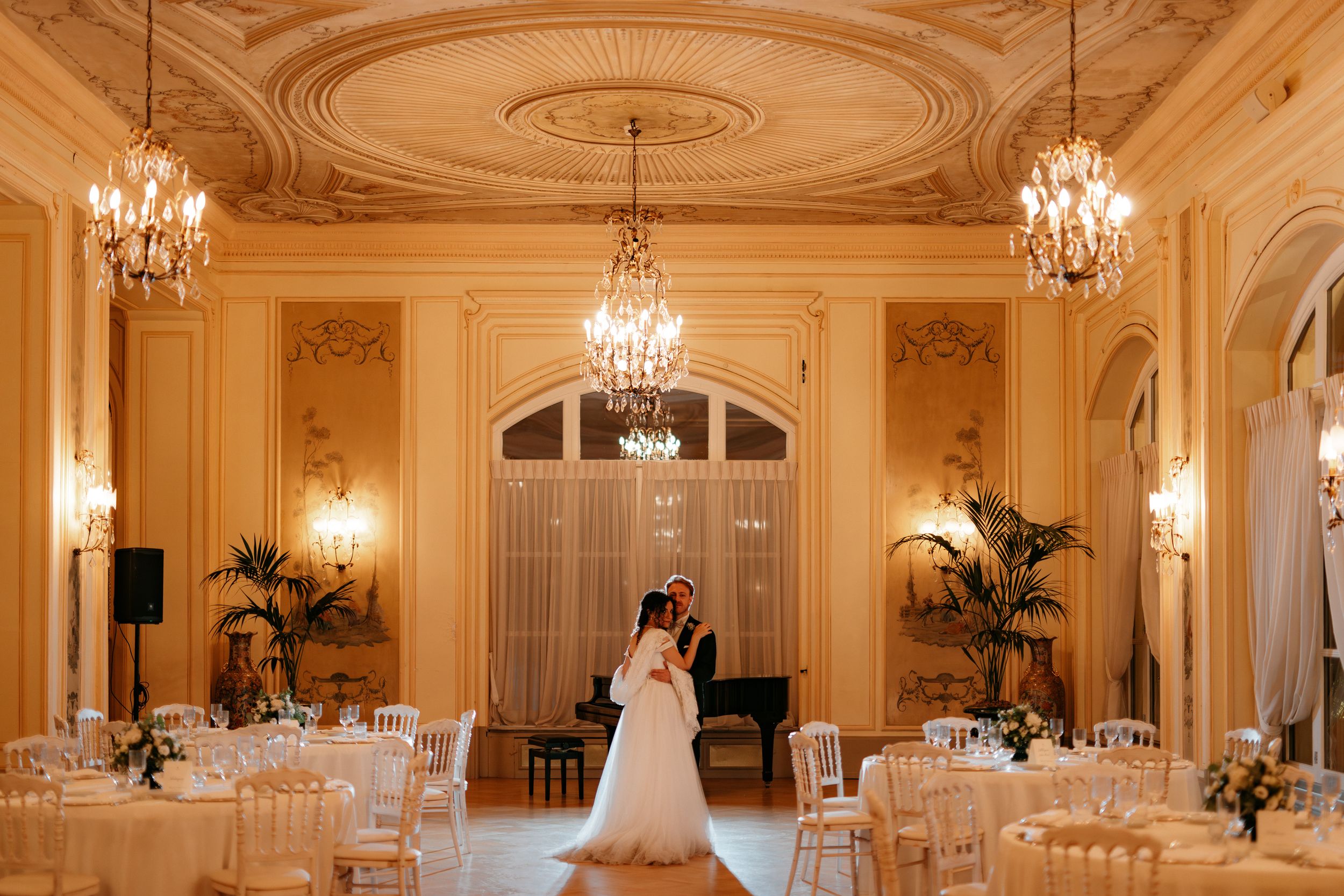 Elegant ballroom with ornate ceiling, crystal chandeliers and white tables set for wedding reception.
