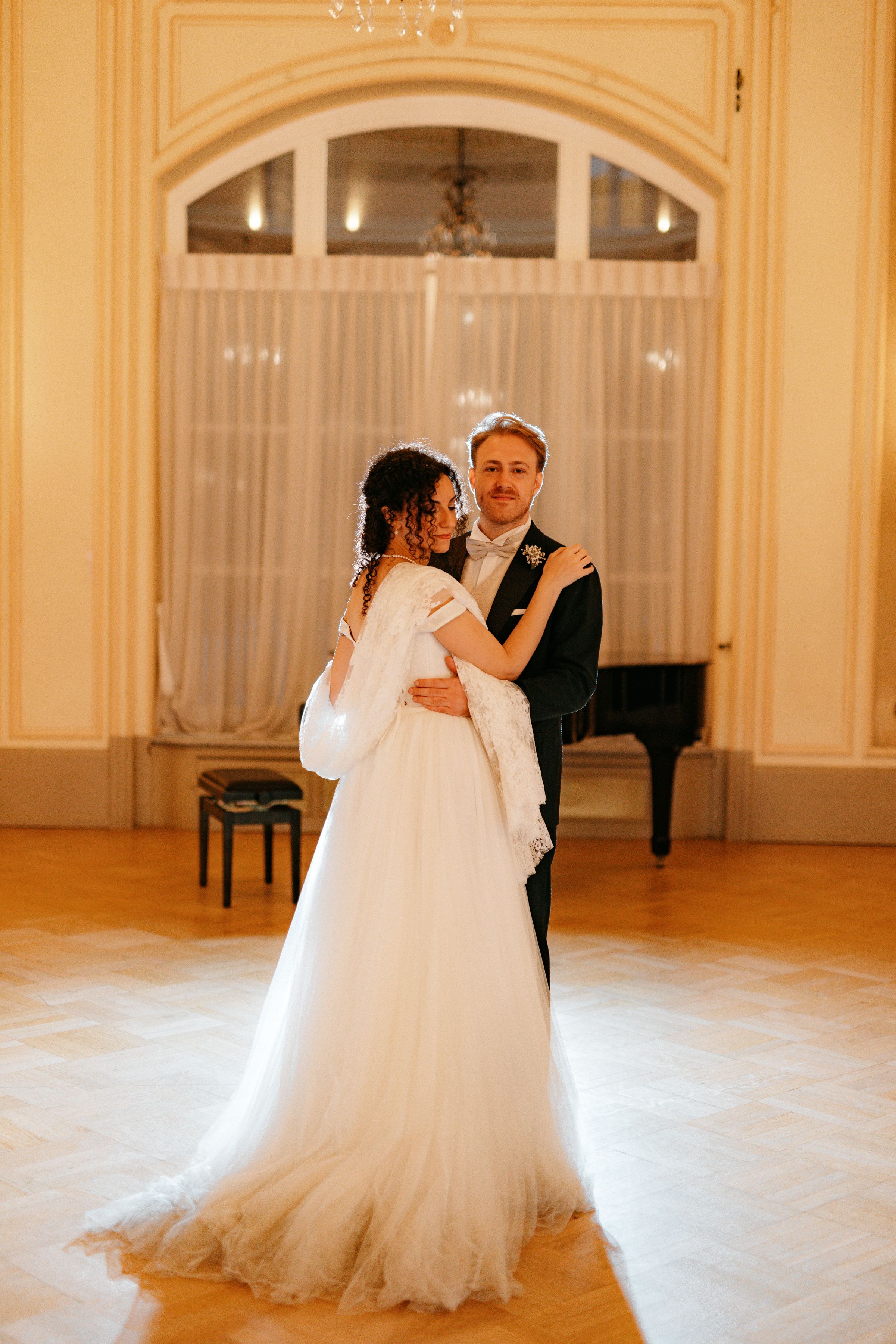 A couple shares their first dance in an elegant ballroom with a grand piano and arched windows in warm lighting.