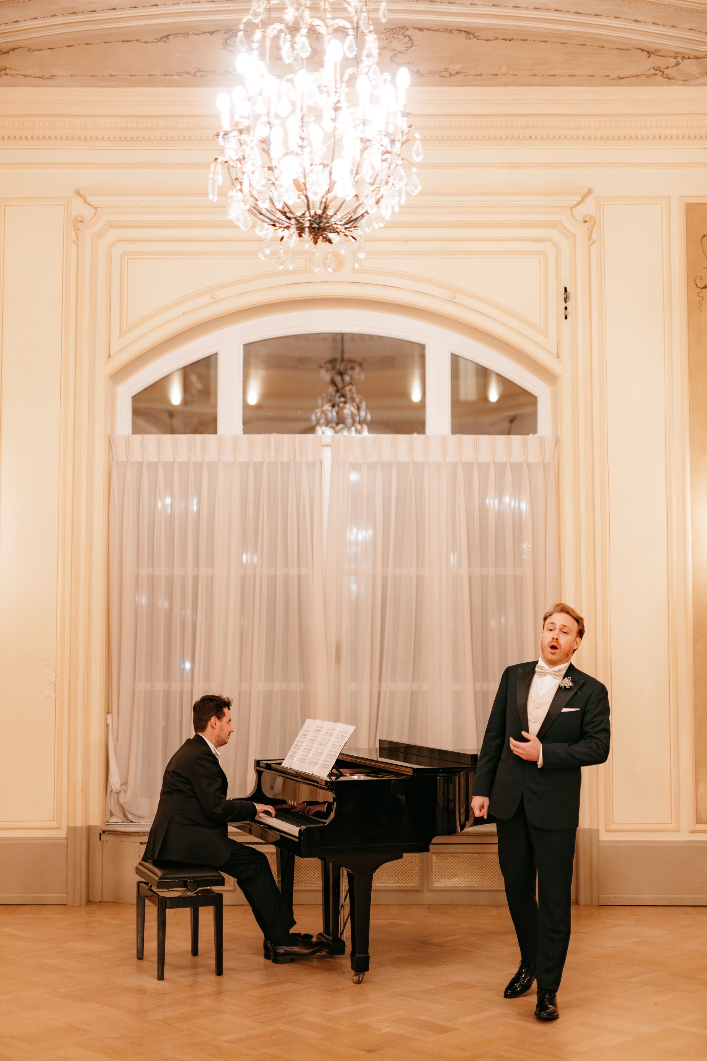 Classical singer performs in elegant white ballroom with crystal chandeliers and grand piano.