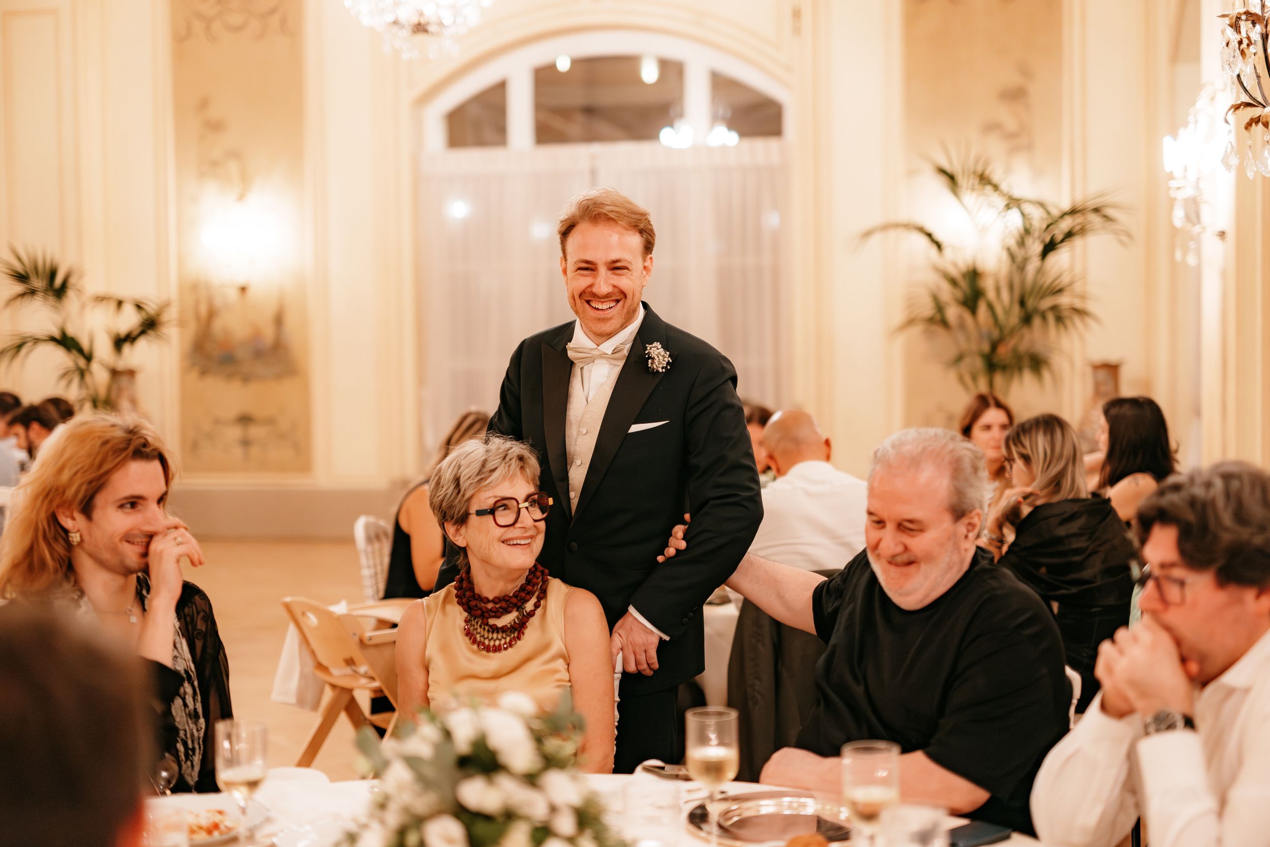 Guests enjoy an elegant dinner reception in a bright white ballroom with ornate chandeliers and floral centerpieces.