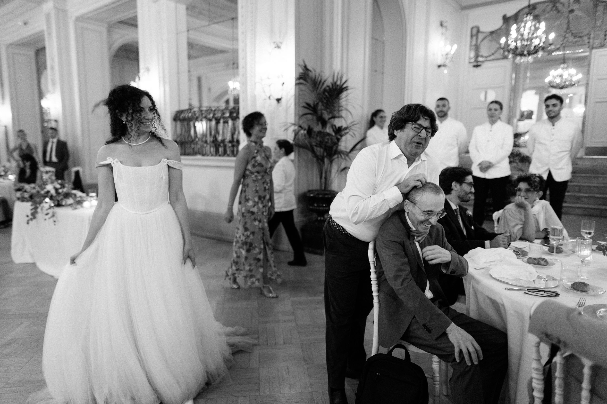 Elegant wedding reception in a grand white ballroom with guests dining at round tables under crystal chandeliers.