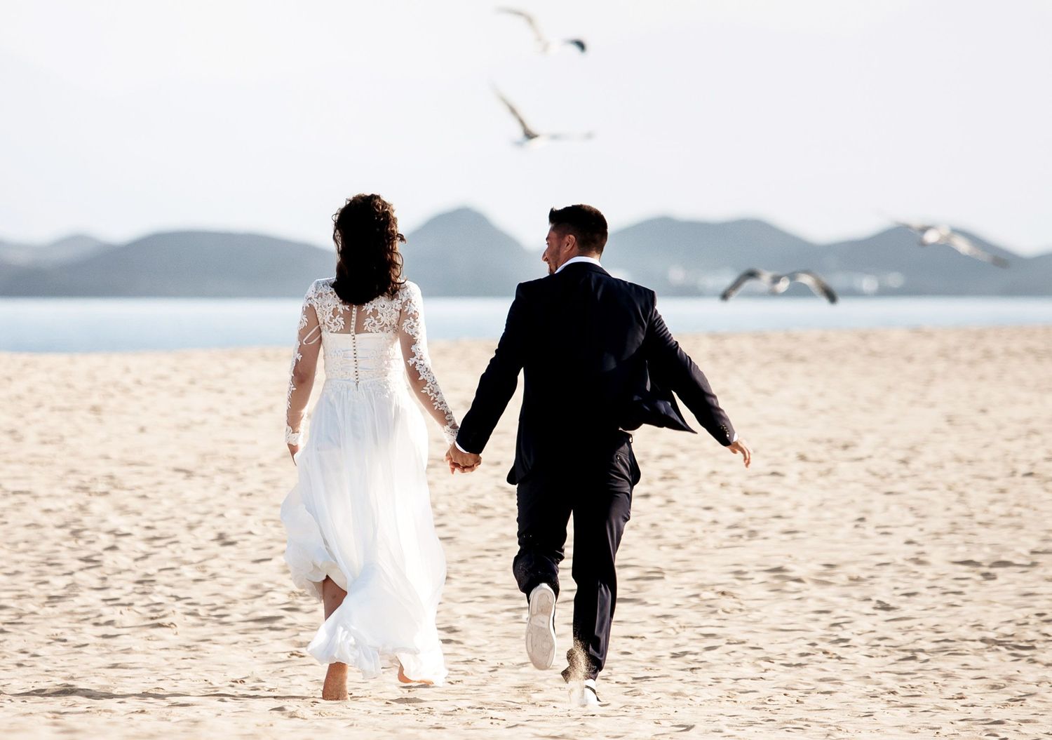 Bride and groom holding hands walking on a sandy beach in Corralejo with seagulls flying overhead and mountains in the background.
