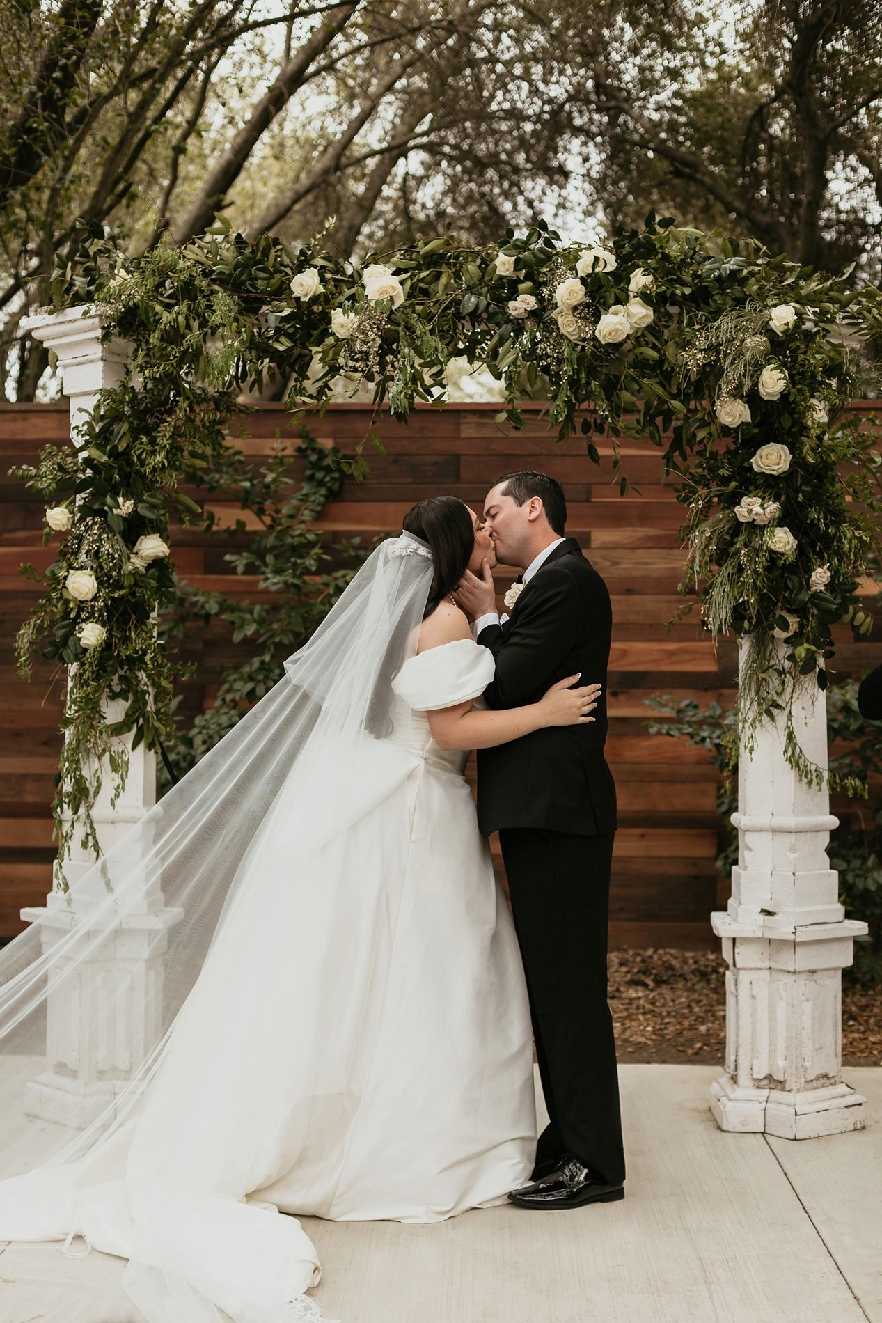 A romantic wedding couple shares a tender kiss against a rustic wooden wall adorned with white floral arrangements.