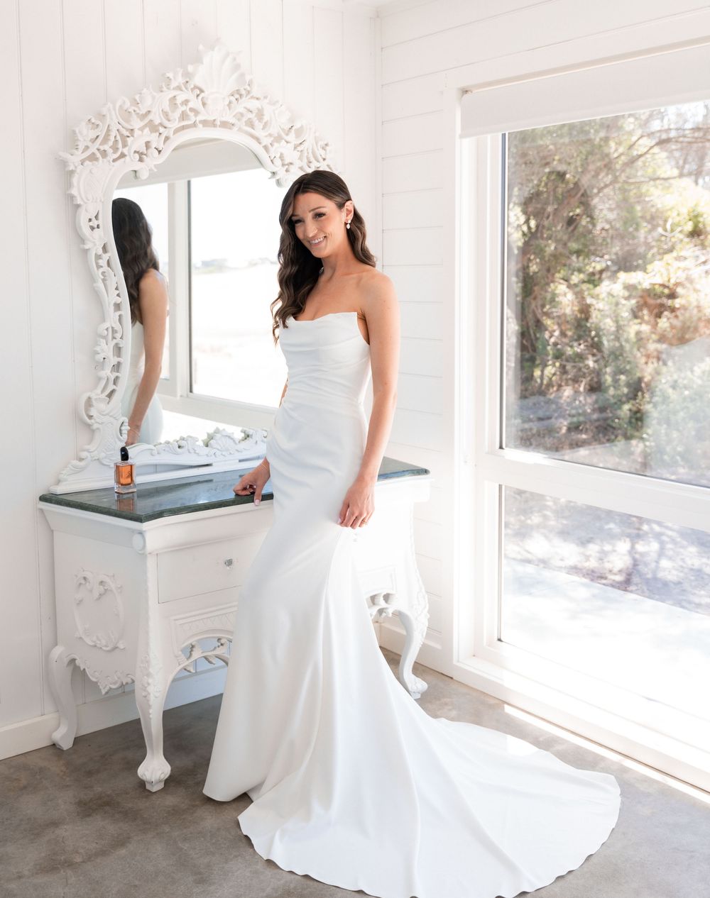 A wedding dress is showcased in a bright, elegant room with white decor and natural light streaming through windows.