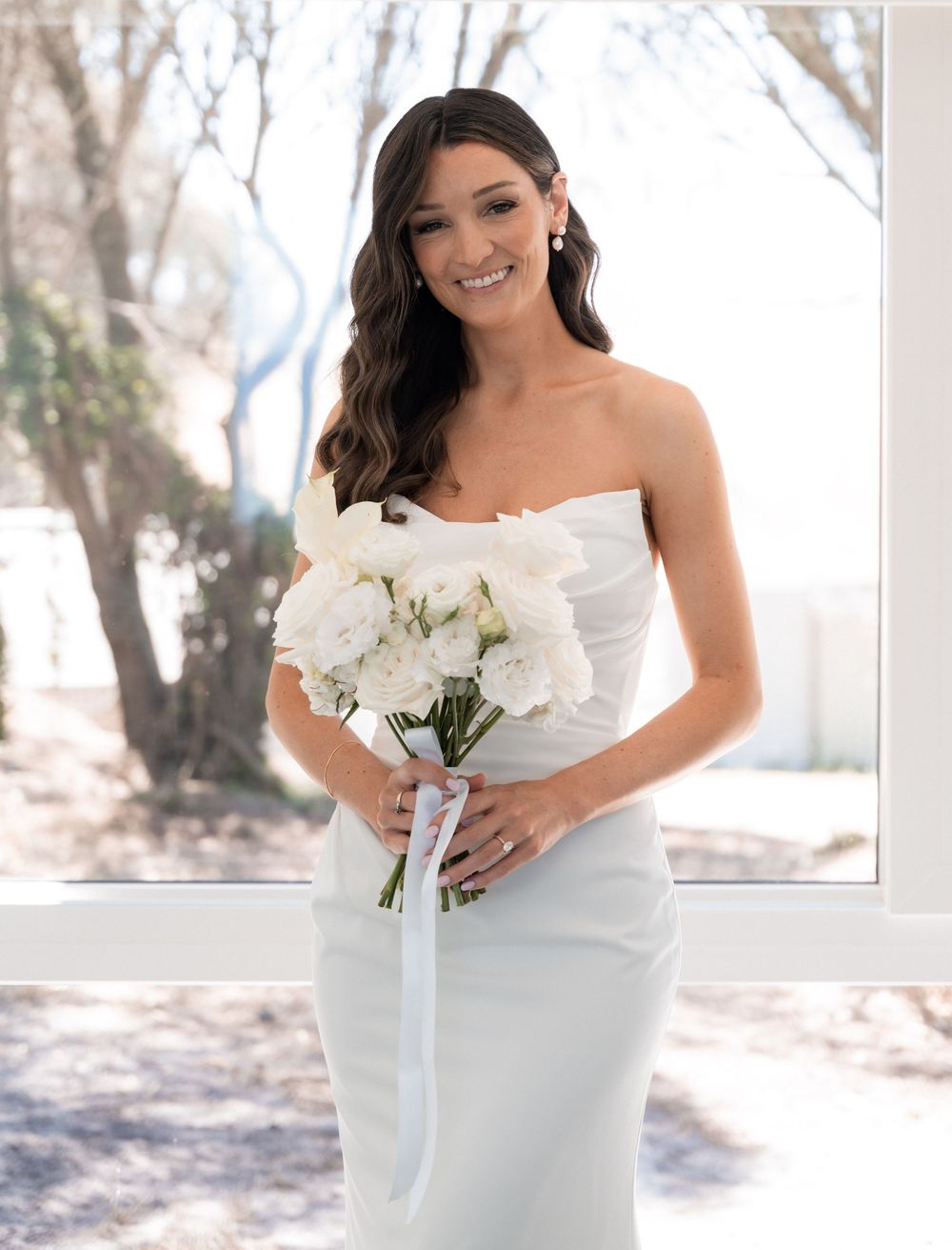 Elegant bride in strapless white gown holds a classic white floral bouquet in natural outdoor setting.