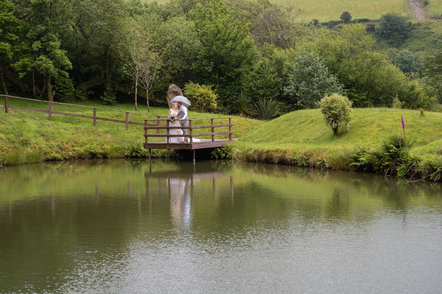 A couple stands under a clear umbrella in the rain during their wedding day celebration.