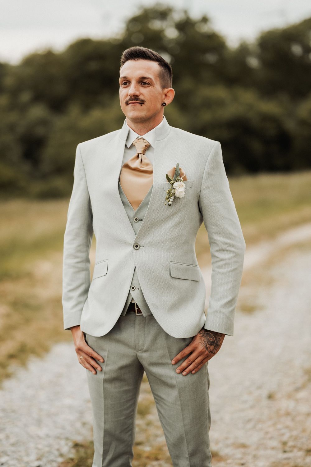 A groom in a light gray suit poses outdoors on a gravel path with trees in the background.