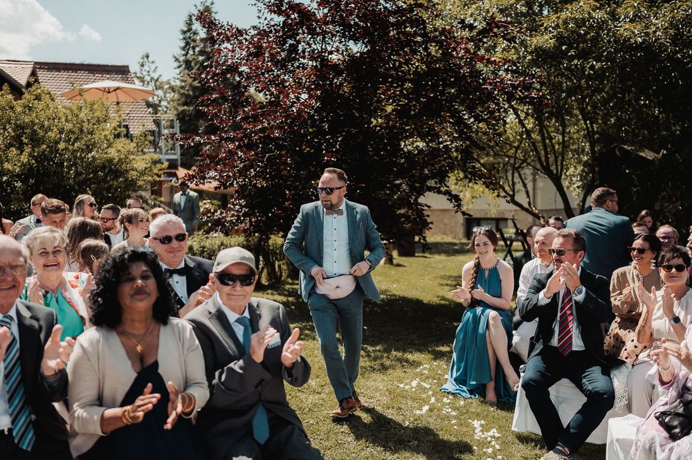 Wedding guests gather outdoors on a sunny day celebrating with bubbles and laughter in a garden setting.