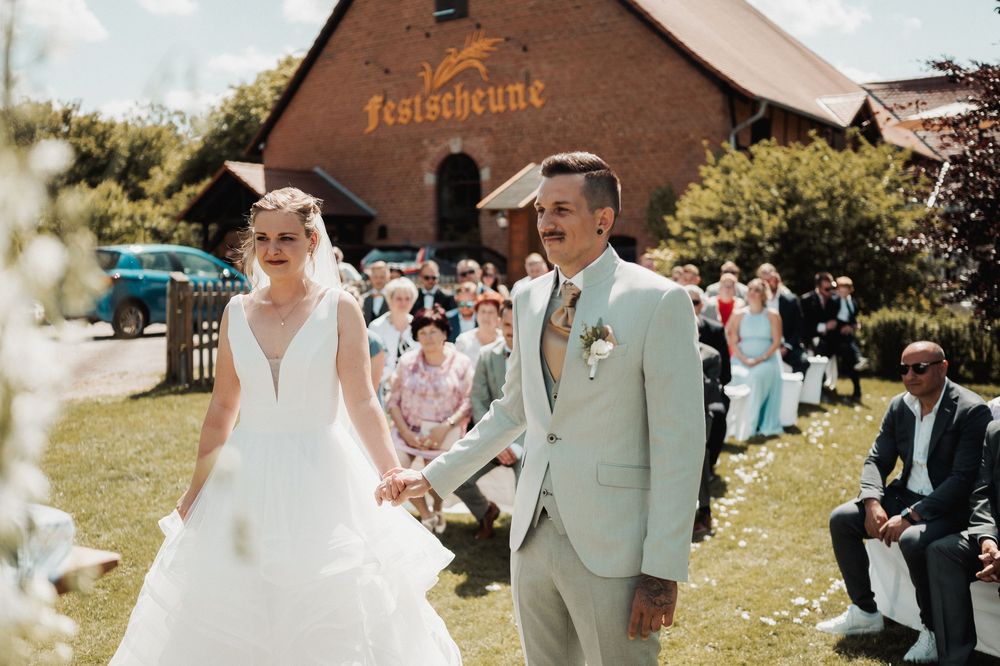A wedding ceremony taking place outdoors with guests seated on white chairs and a rustic building in the background.