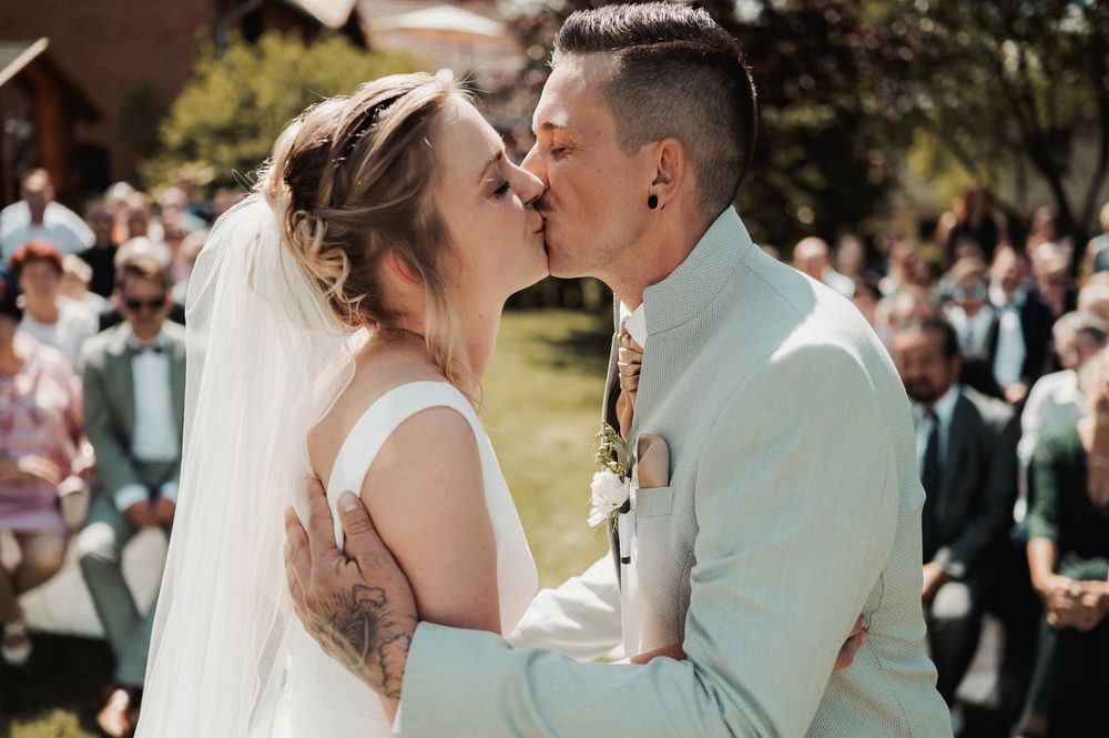 A romantic wedding kiss between newlyweds in white attire surrounded by guests in an outdoor garden setting.