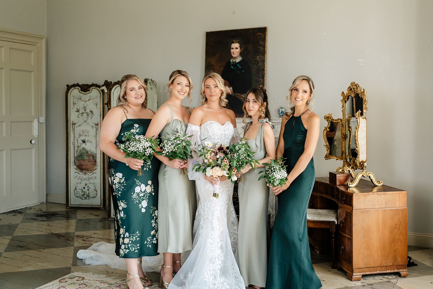 Bridesmaids in elegant green dresses pose together holding bouquets in a vintage-style room with antique decor.