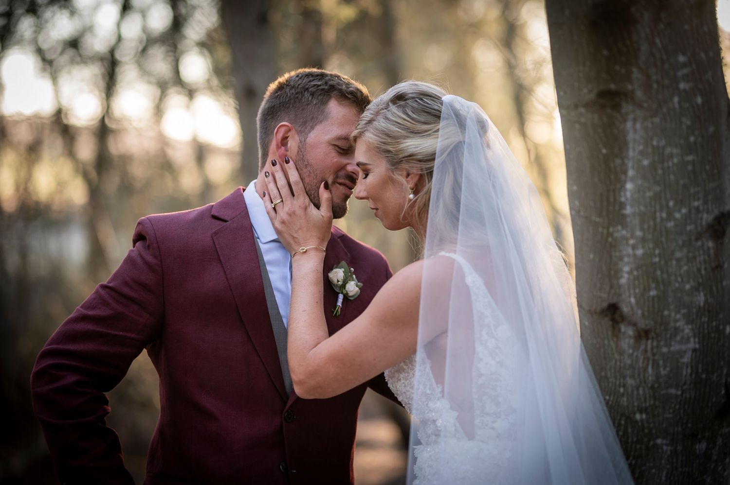 Bride and groom share an intimate moment outdoors, touching foreheads, groom in burgundy suit, bride in white gown with veil.