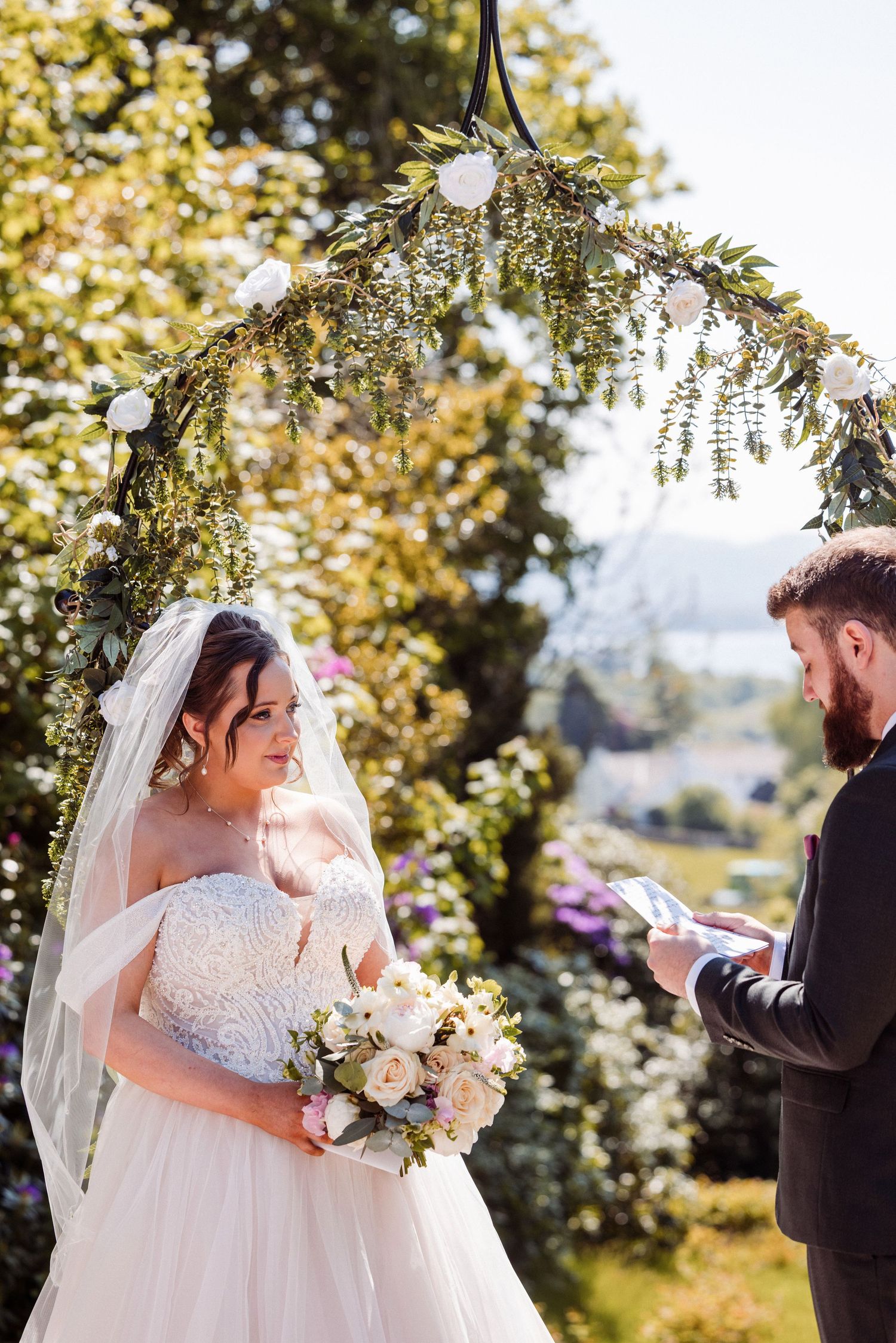 Bride and groom share emotional first look moment in garden setting with sunlit trees in background.